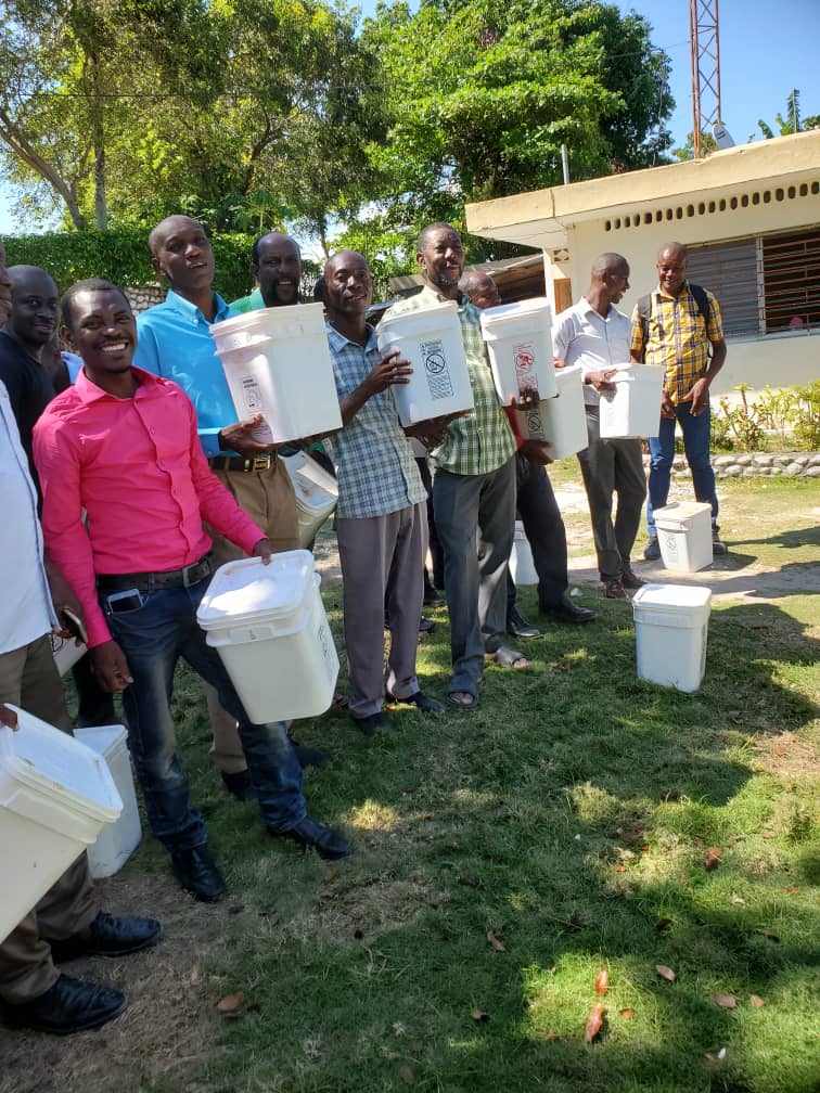 Group of men holding white containers, standing outdoors in a green area, showcasing community engagement and distribution efforts.