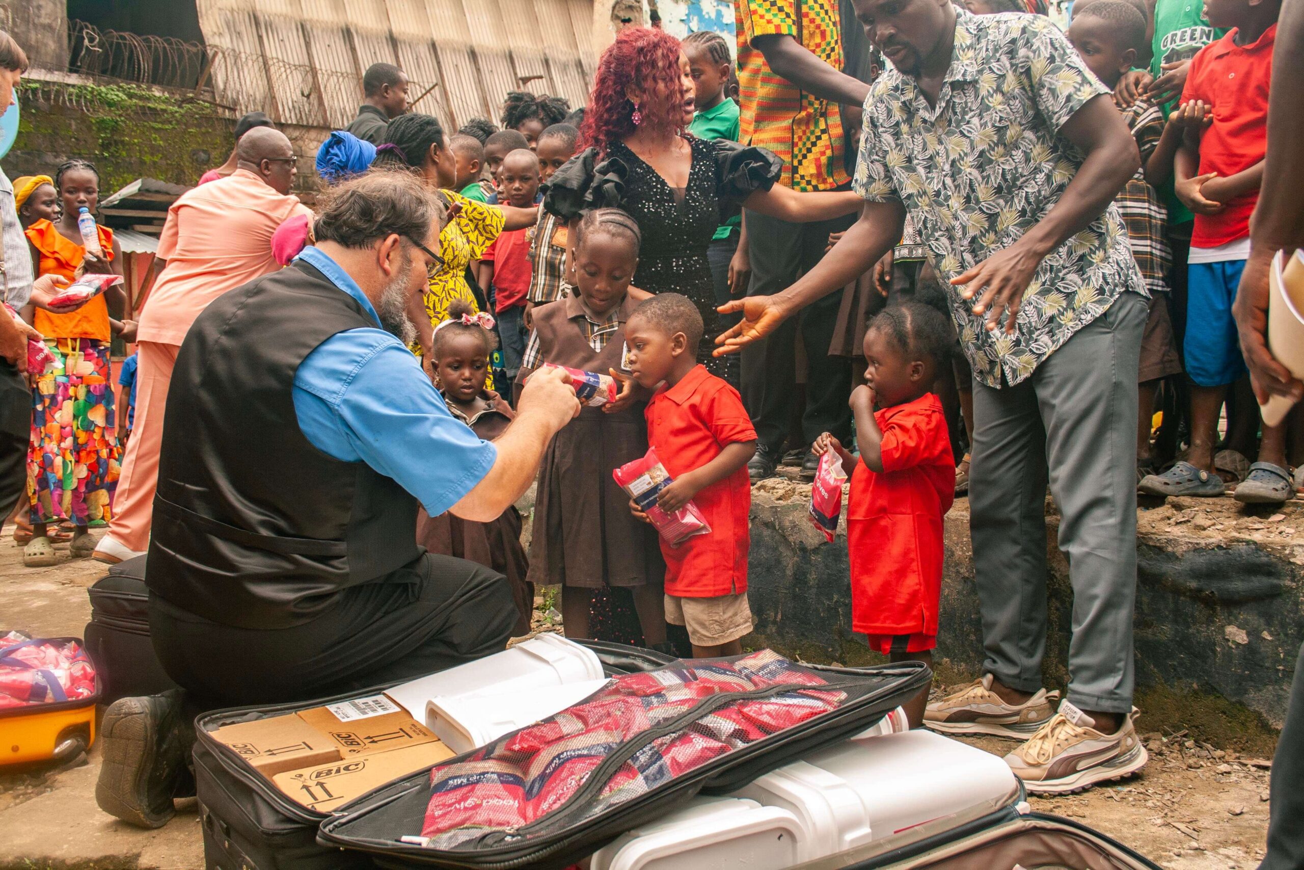 A group of children eagerly receive snacks and drinks from volunteers in a community outreach event, with adults assisting and engaging in a vibrant urban setting.