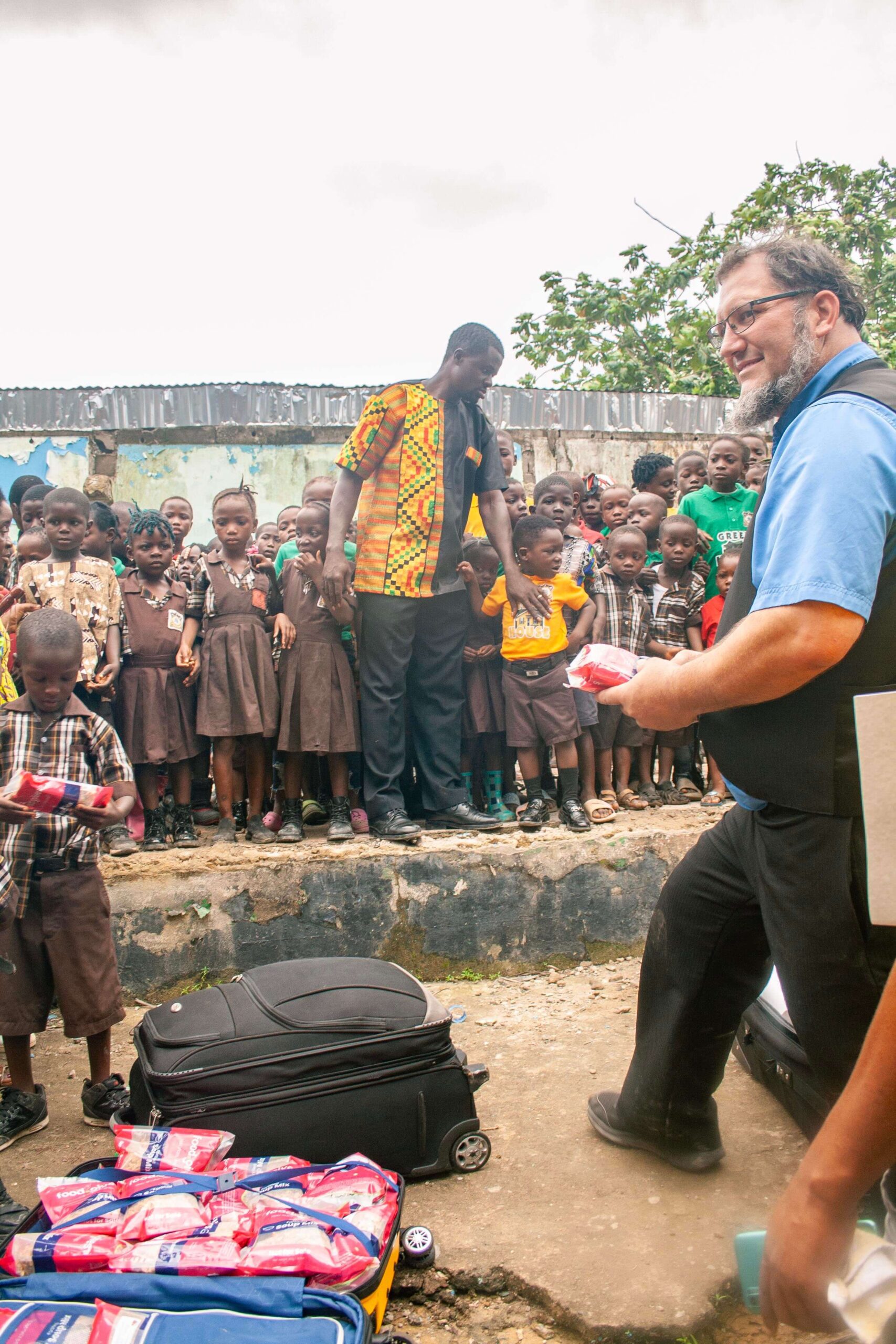 Children in school uniforms gather excitedly around adults distributing supplies in an outdoor setting, with bags and suitcases visible on the ground. The scene showcases community engagement and support for education.