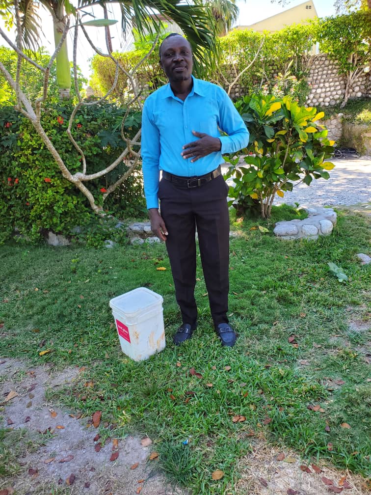 Man in a blue shirt standing on grass next to a white container, surrounded by lush greenery and tropical plants. The scene captures a vibrant outdoor setting, ideal for showcasing gardening or landscaping themes.