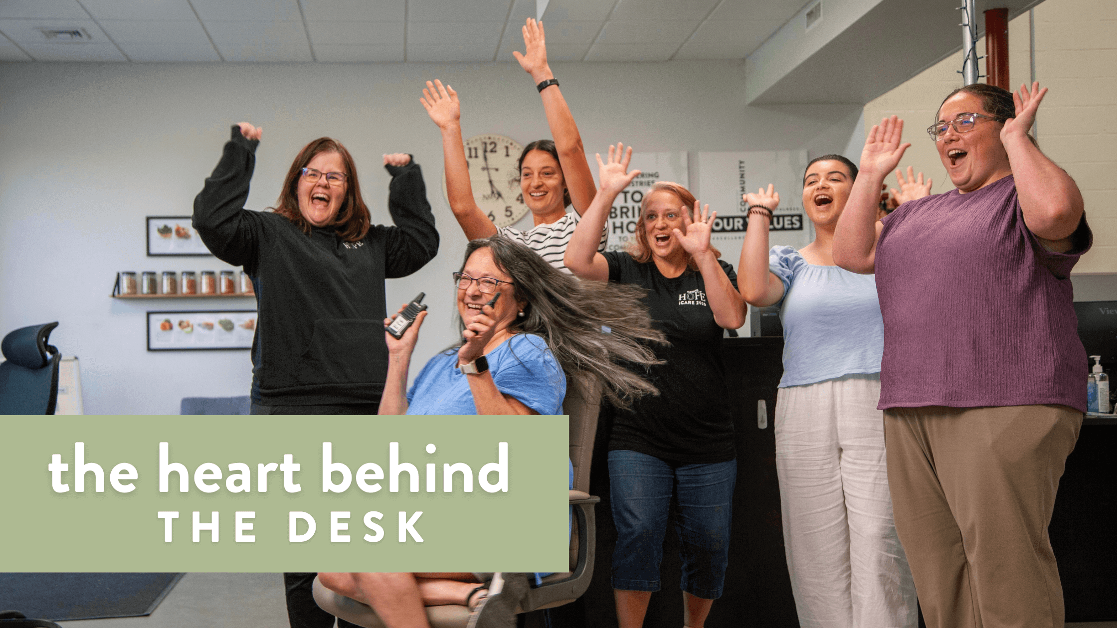 Group of cheerful women celebrating together in an office setting, showcasing teamwork and positivity. The image highlights a sense of community and joy among colleagues, with the phrase "the heart behind the desk" prominently displayed.