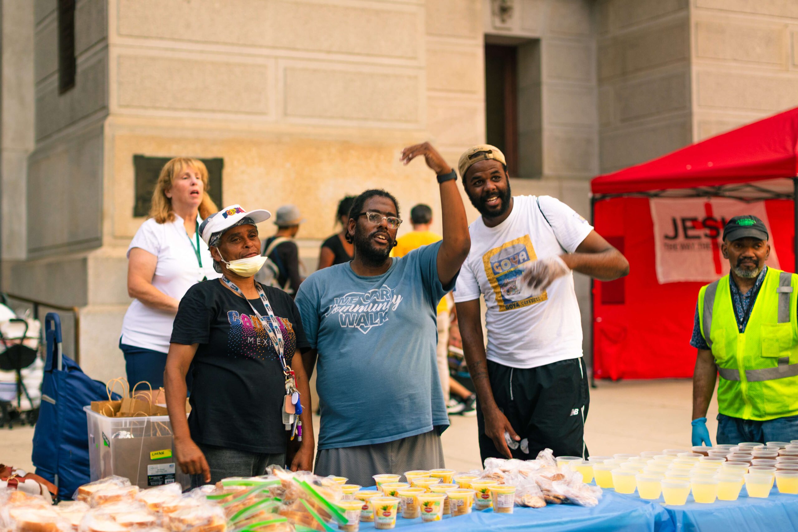 Community volunteers distribute food and drinks at a local event, showcasing a diverse group of people engaging with attendees. The scene features a table filled with packaged meals and refreshments, emphasizing community support and outreach efforts.