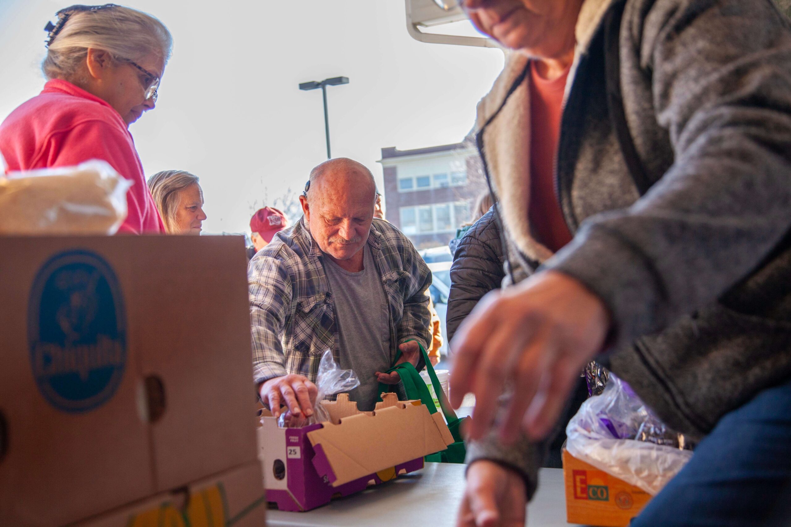 Community members participating in a food distribution event, with individuals sorting through boxes and gathering items at a table. The scene captures the spirit of collaboration and support among attendees.
