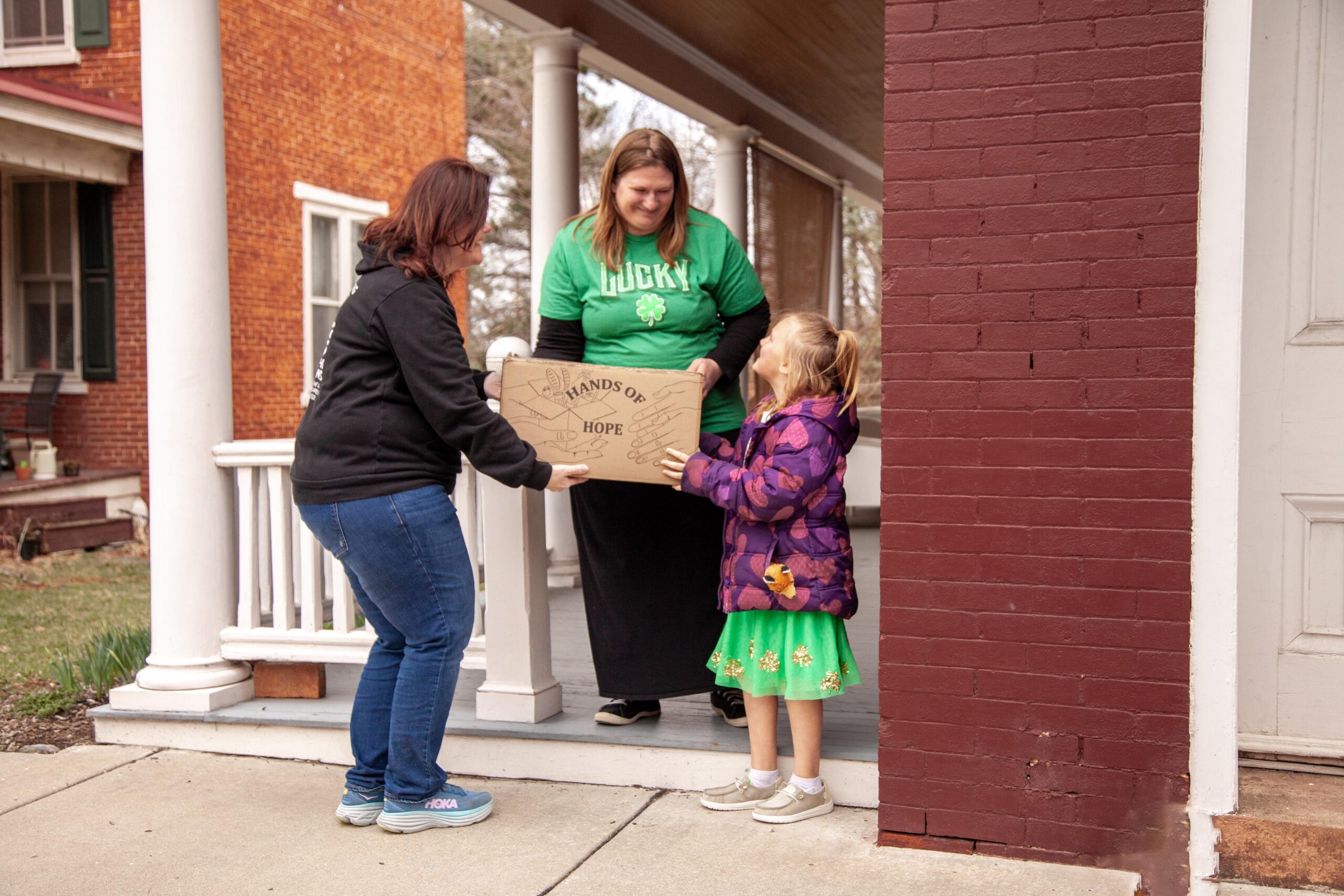 Community members participate in a charity event, delivering a "Hands of Hope" box to a family. The scene features two women and a young girl on a porch, promoting kindness and support within the neighborhood.