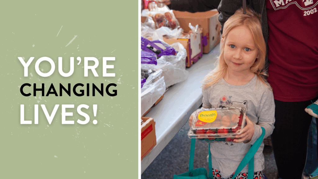 Young girl holding a container of strawberries at a food distribution event, surrounded by boxes of fresh produce, highlighting community support and healthy food access. With a message you're changing lives!