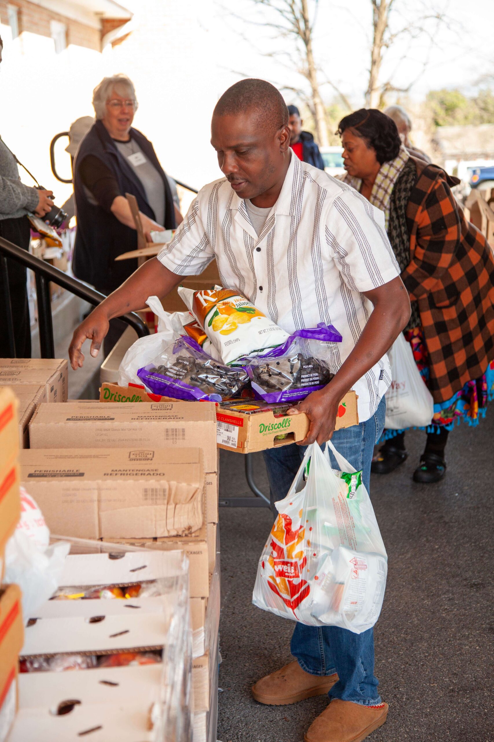 A man carrying a box of food items, including snacks and fruits, while participating in a community food distribution event. In the background, several volunteers assist with the distribution process, highlighting community support and engagement.