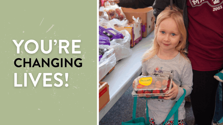 Young girl holding a container of strawberries at a food distribution event, surrounded by boxes of fresh produce, highlighting community support and healthy food access. With a message you're changing lives!