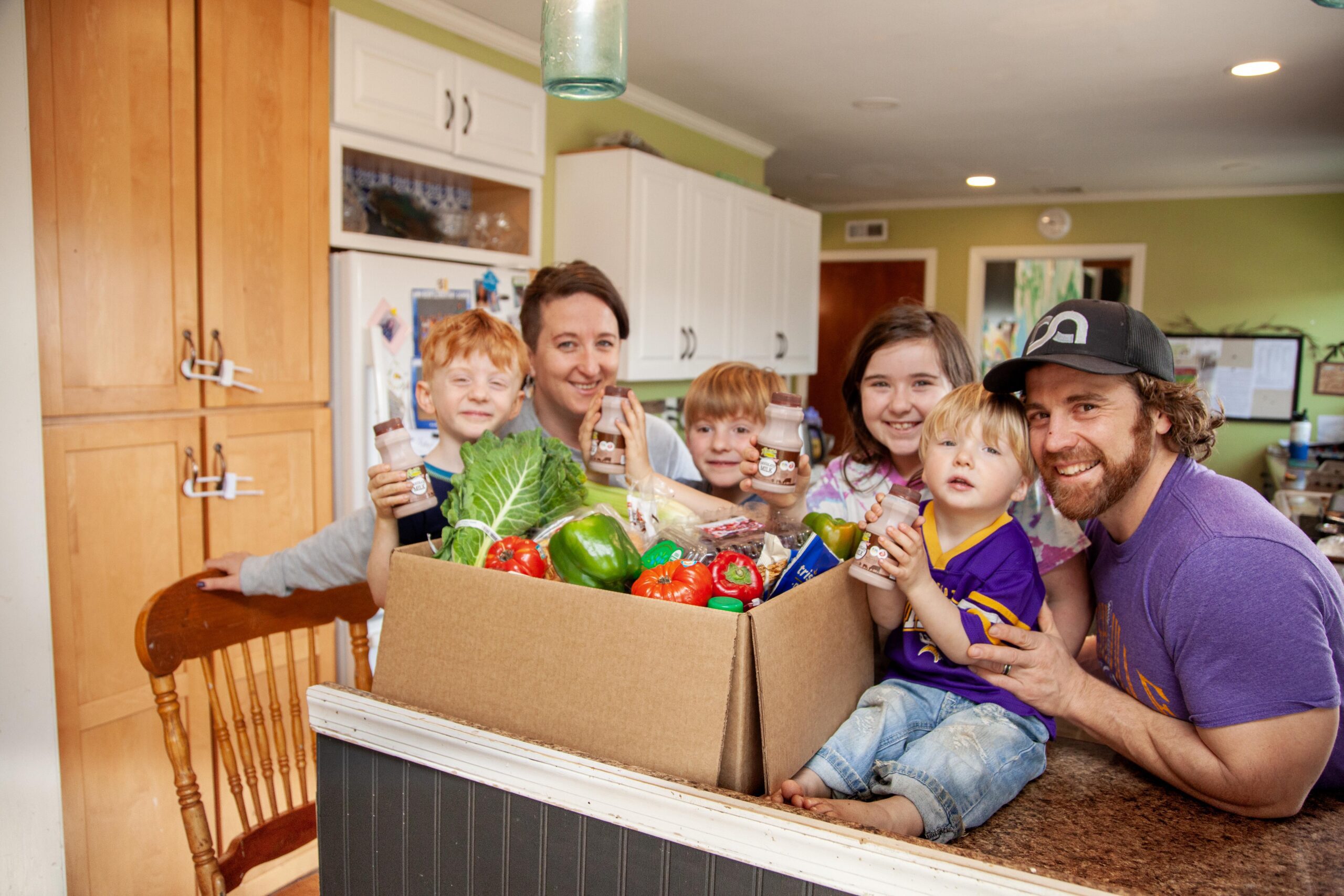 Family enjoying a healthy lifestyle by posing with a large box of fresh groceries, including vegetables and drinks, in a bright kitchen setting.