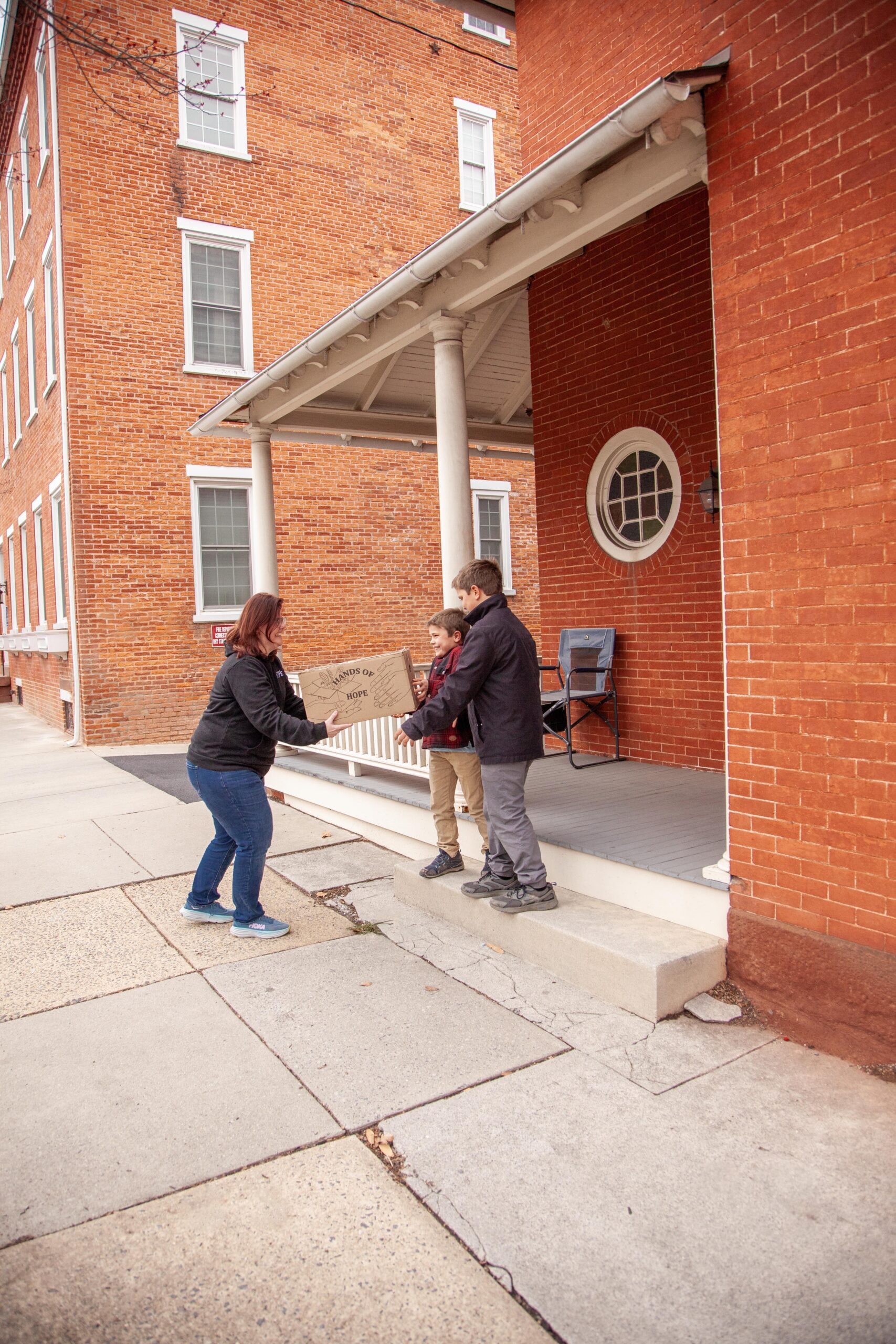 A woman hands a box labeled "Hands of Hope" to two boys on the steps of a brick building, showcasing a moment of community engagement and support.
