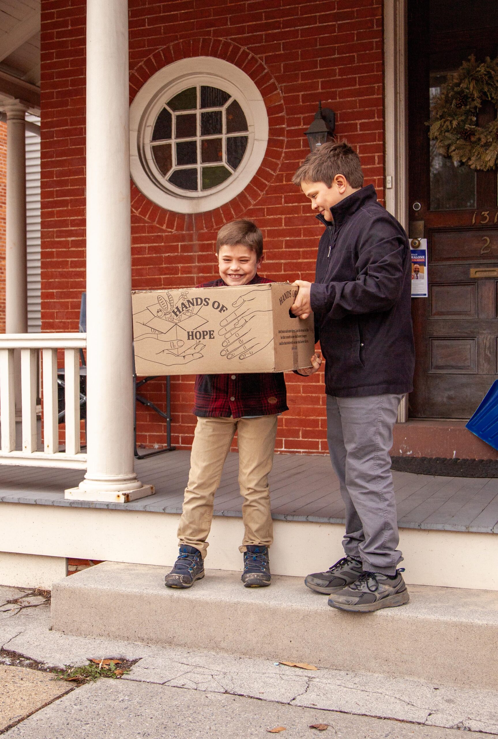 Two young boys smiling while holding a "Hands of Hope" donation box on a front porch, showcasing community support and charity efforts. The background features a red brick wall and a decorative door, emphasizing a welcoming home environment.
