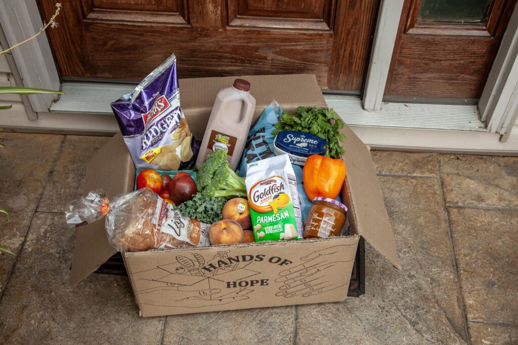 Cardboard box labeled "Hands of Hope" filled with assorted groceries including chips, chocolate milk, fresh fruits like apples and peaches, vegetables like broccoli and bell pepper, and various snacks, placed at a doorway.