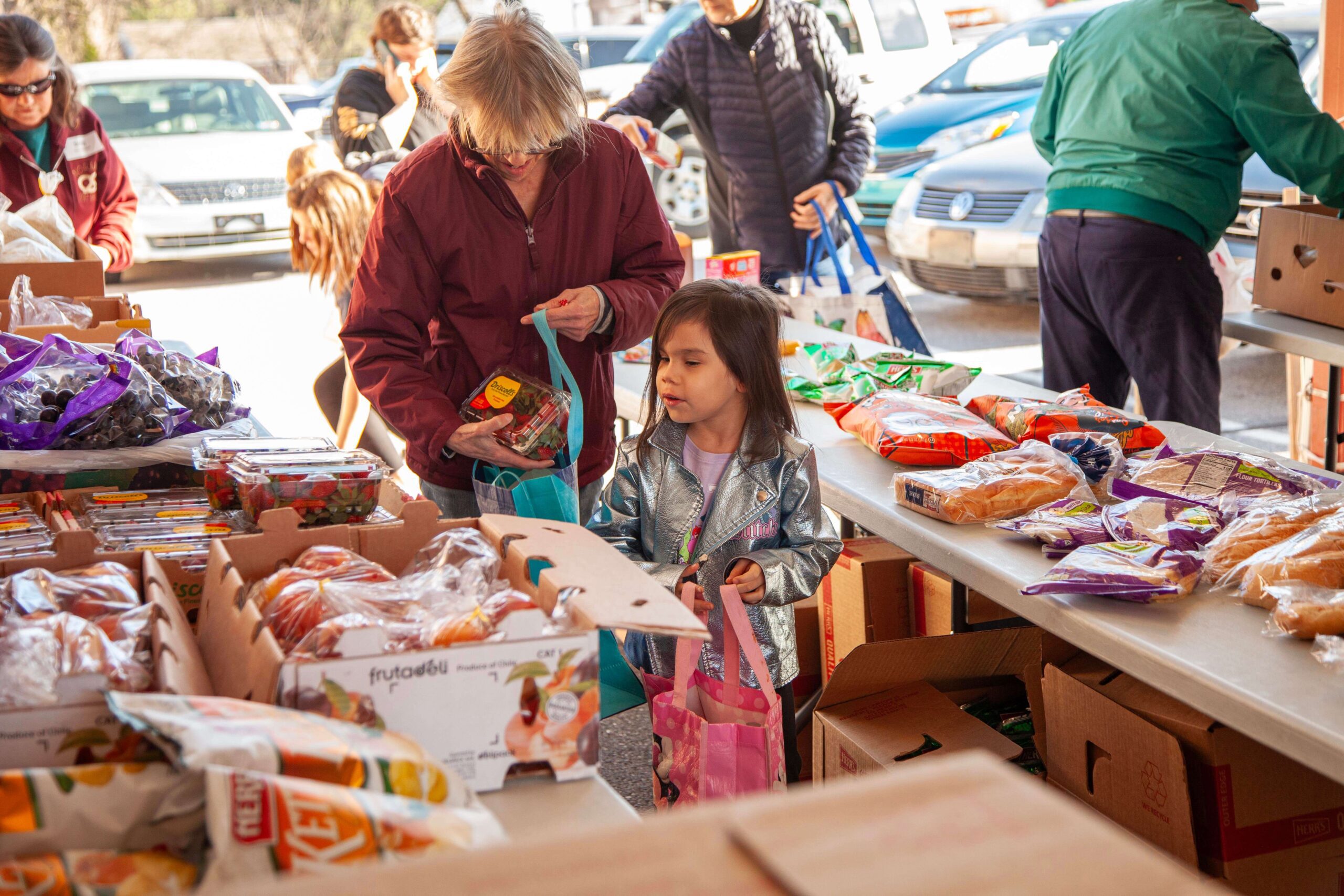 A woman and a young girl select food items from a table filled with fresh produce and packaged goods at a community food distribution event. The scene captures the importance of access to healthy food in a supportive environment.