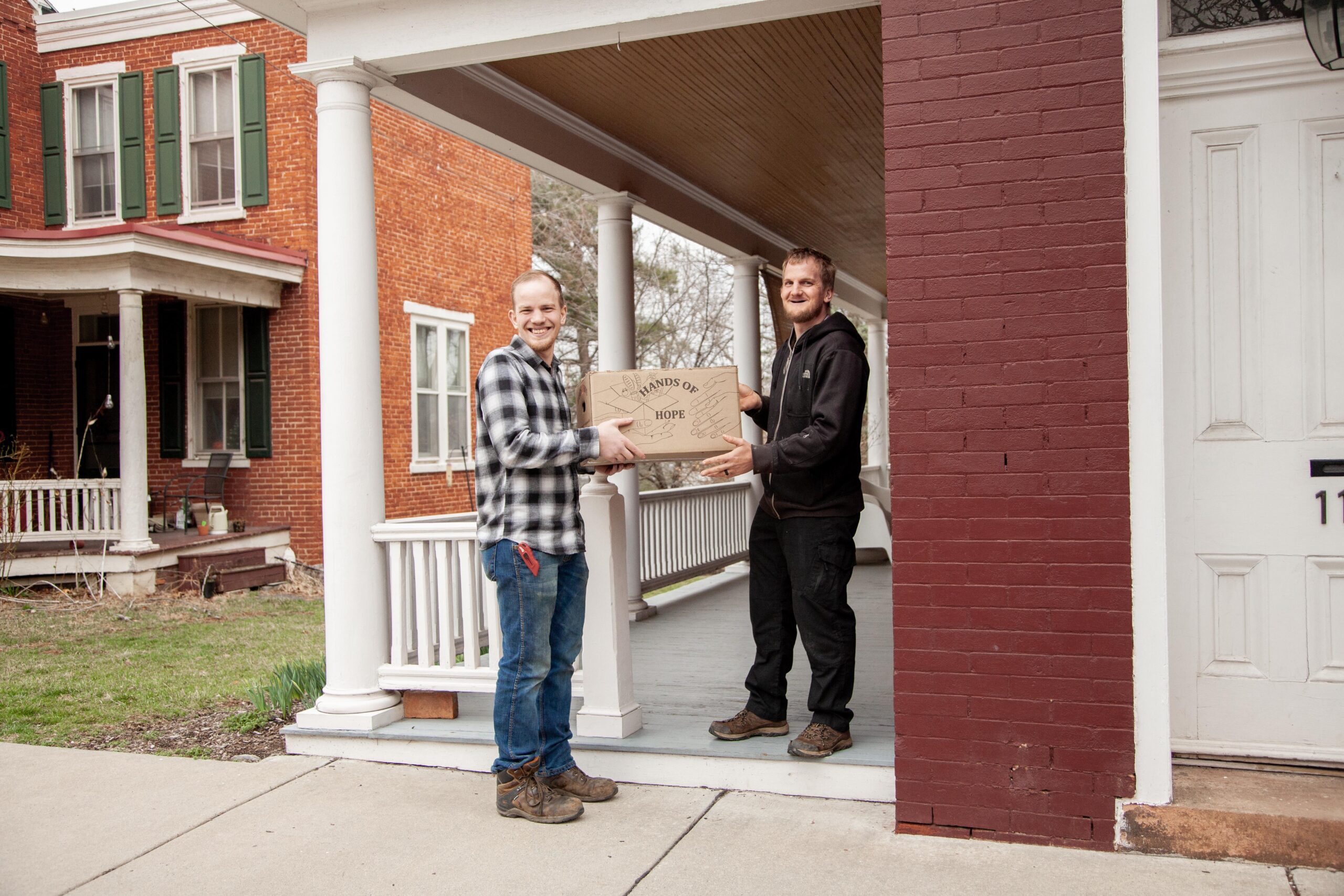 Two men are smiling and exchanging a box labeled 'Hands of Hope' on a porch in front of a brick house. The scene captures a moment of community support and delivery service.