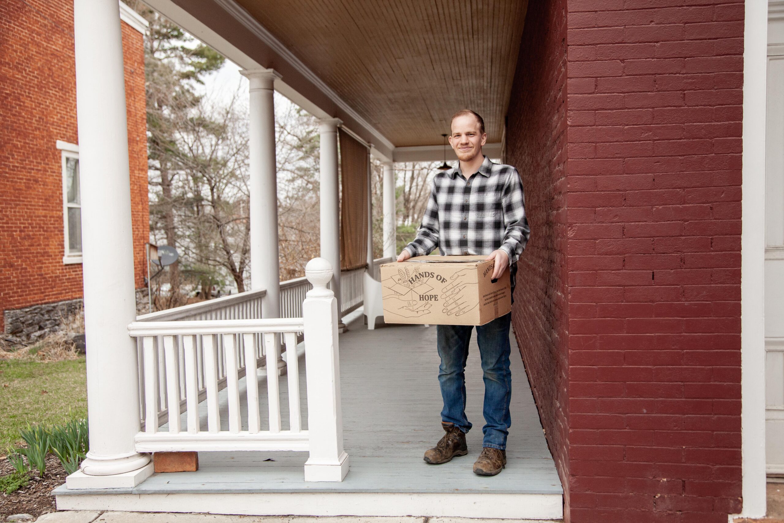 Man holding a box labeled "Hands of Hope" while standing on a porch, showcasing community support and outreach efforts. The background features a mix of brick and wooden structures, emphasizing a welcoming neighborhood environment.