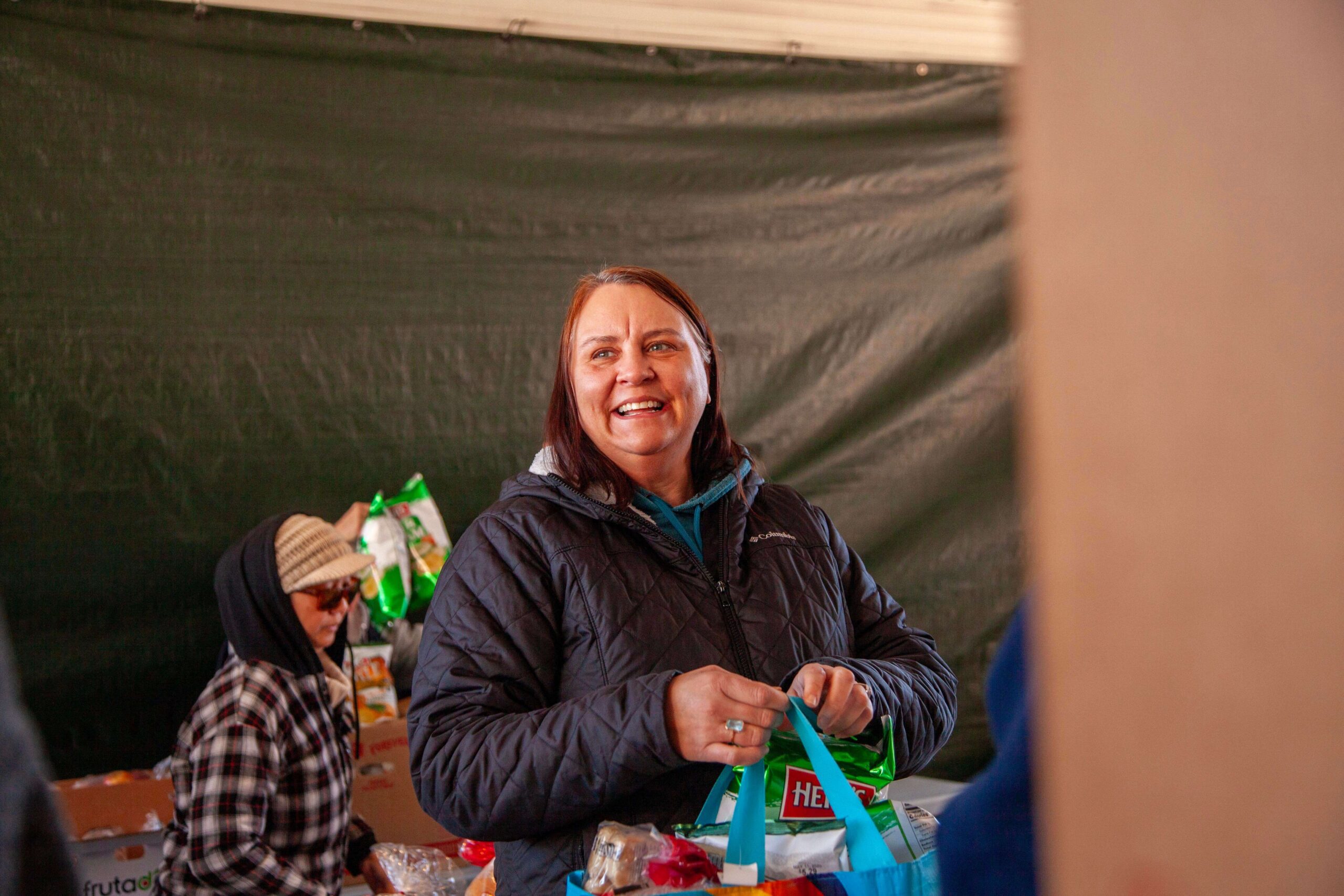 Smiling woman in a black jacket holding a blue tote bag filled with groceries at a food distribution event, with a green backdrop and other volunteers in the background.