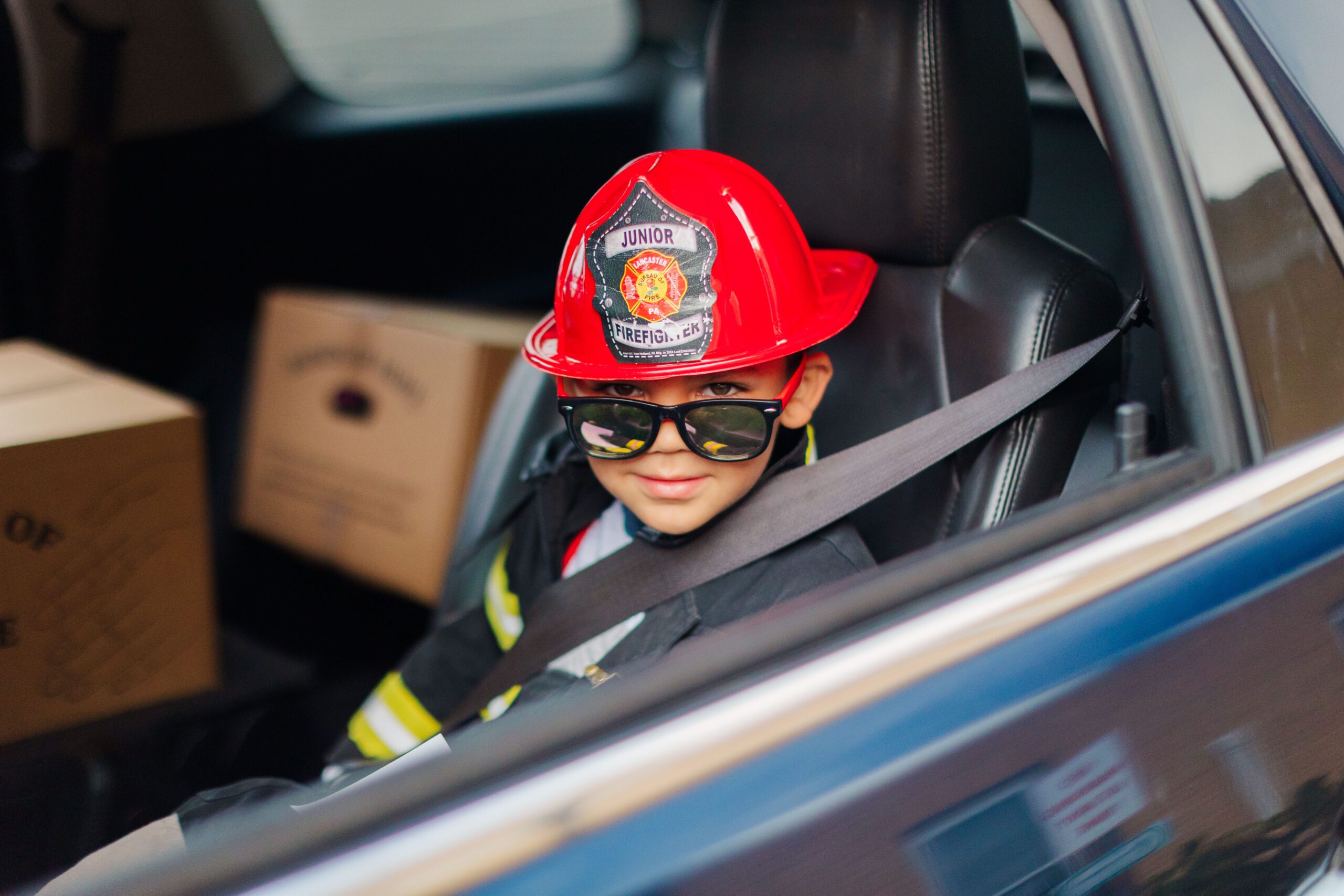 Young boy wearing a red firefighter hat and sunglasses, sitting in the driver's seat of a car, with cardboard boxes visible in the background.