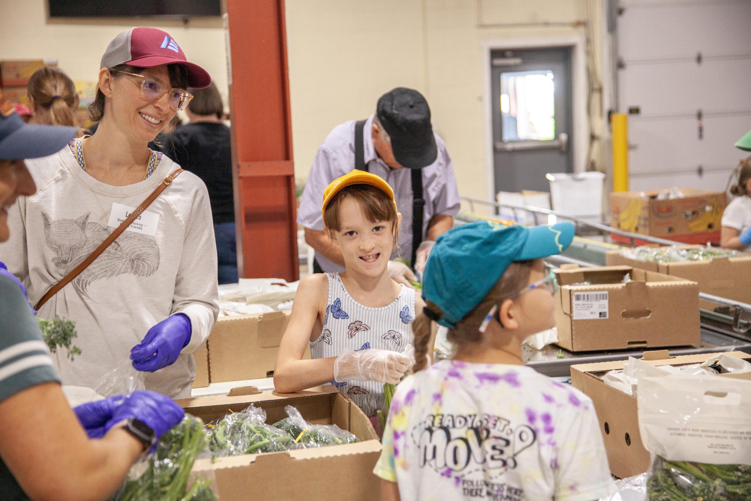 A group of volunteers, including children and adults, participate in a food distribution event, sorting and packaging fresh produce in a community center. The scene highlights teamwork and community involvement in supporting local food initiatives.