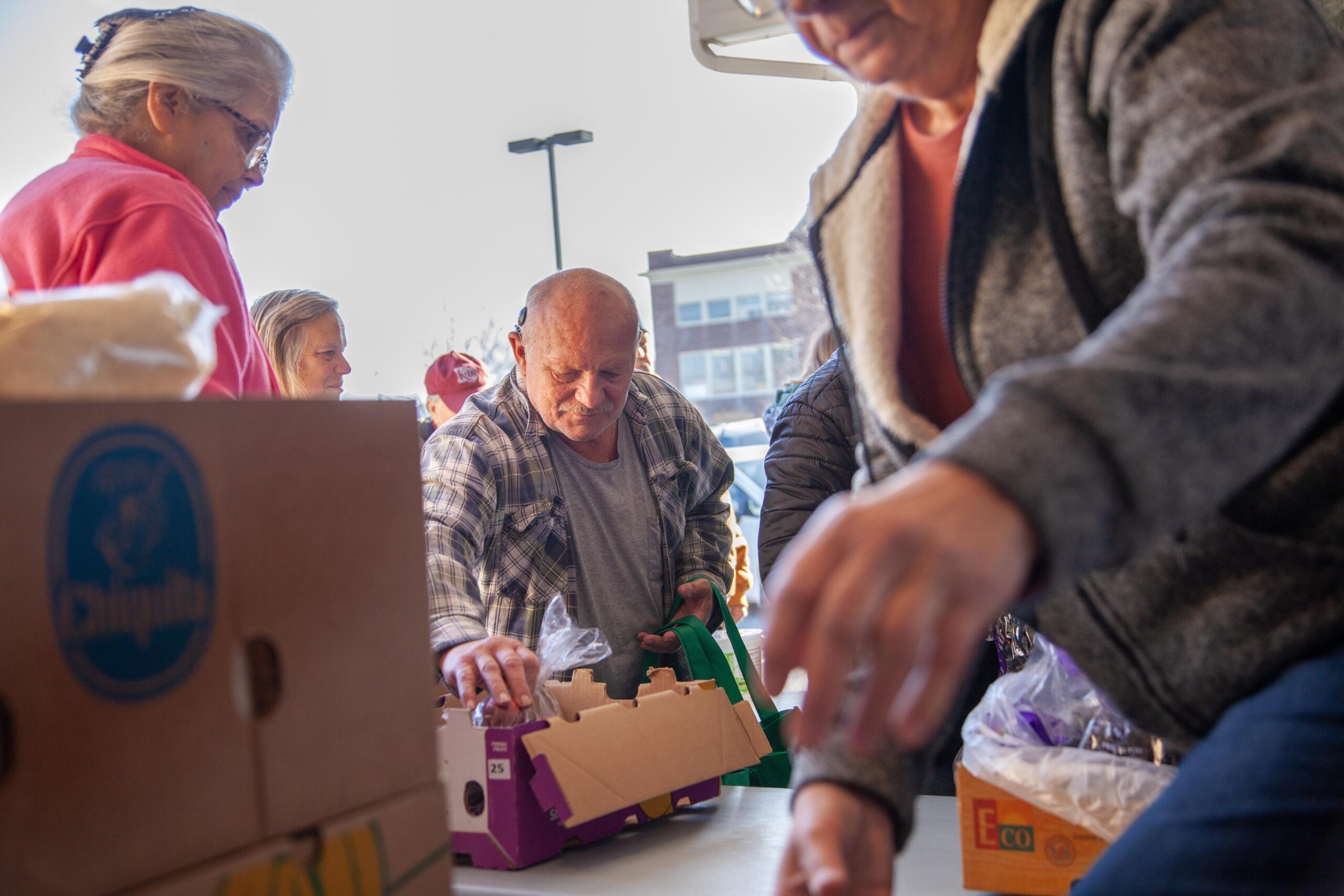 Community members participating in a food distribution event, selecting items from boxes on a table. The scene showcases diverse individuals engaged in the process, highlighting the importance of community support and food assistance programs.