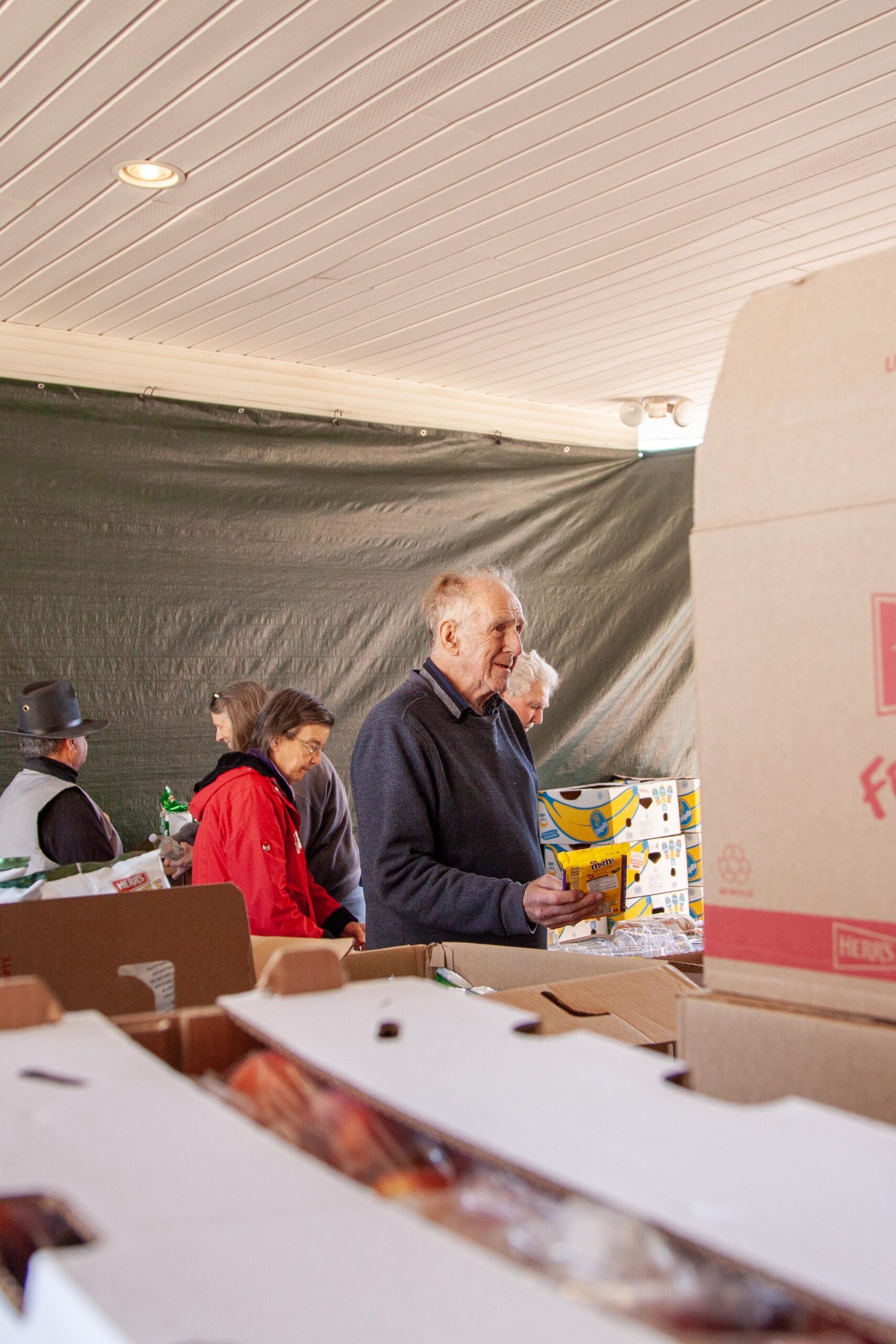 Elderly man shopping for groceries at a community food distribution event, surrounded by volunteers and various food boxes. The scene captures a warm atmosphere of community support and assistance.
