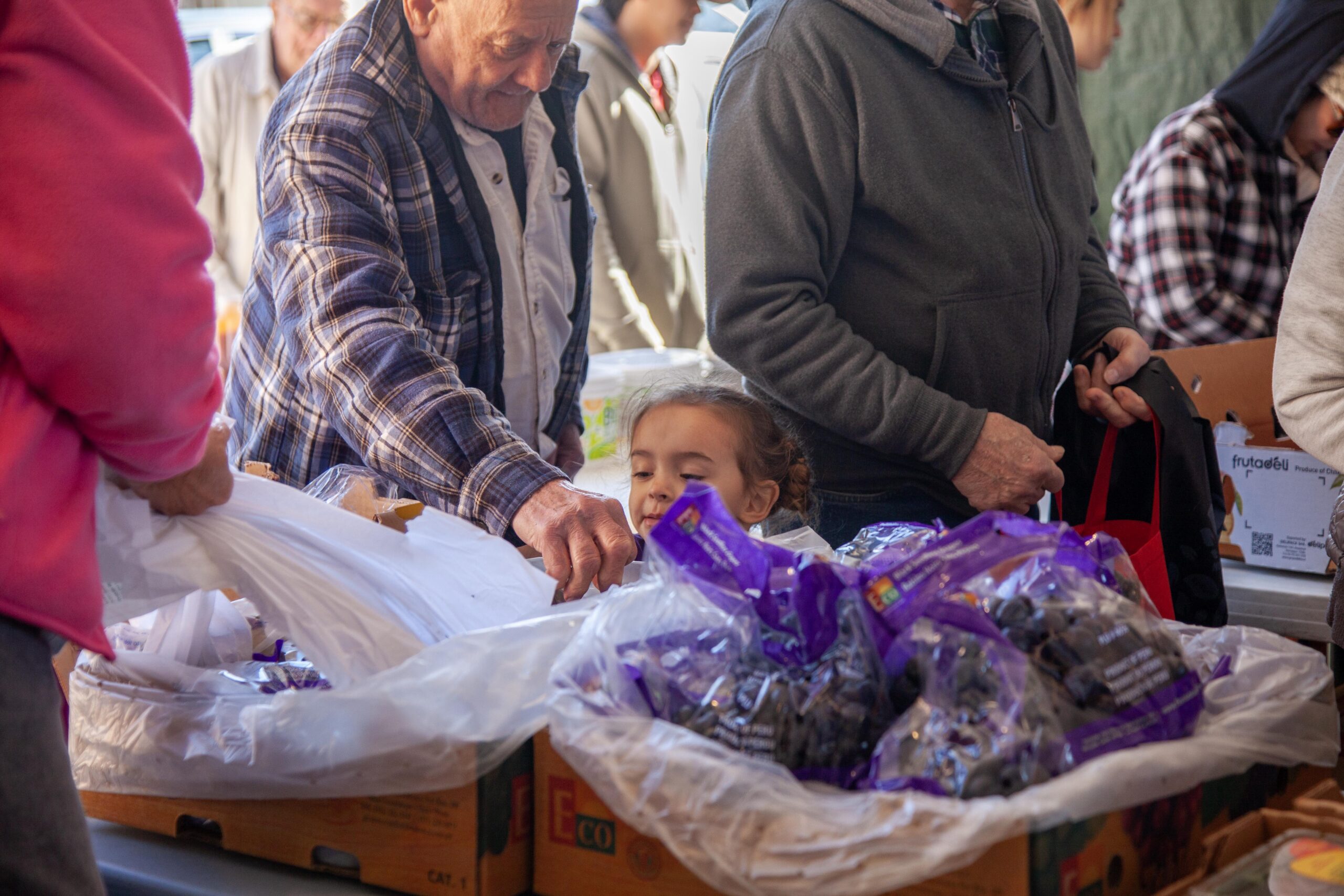 A community member helps a young girl select fresh produce at a local market, surrounded by boxes of fruits and vegetables. The image captures the vibrant atmosphere of a food distribution event, highlighting intergenerational interaction and community engagement.