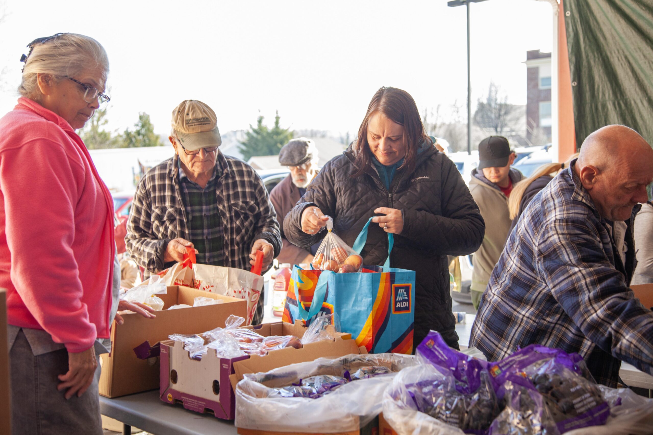 Community members participate in a food distribution event, selecting fresh produce and packaged goods from tables filled with various items, highlighting local support and collaboration.