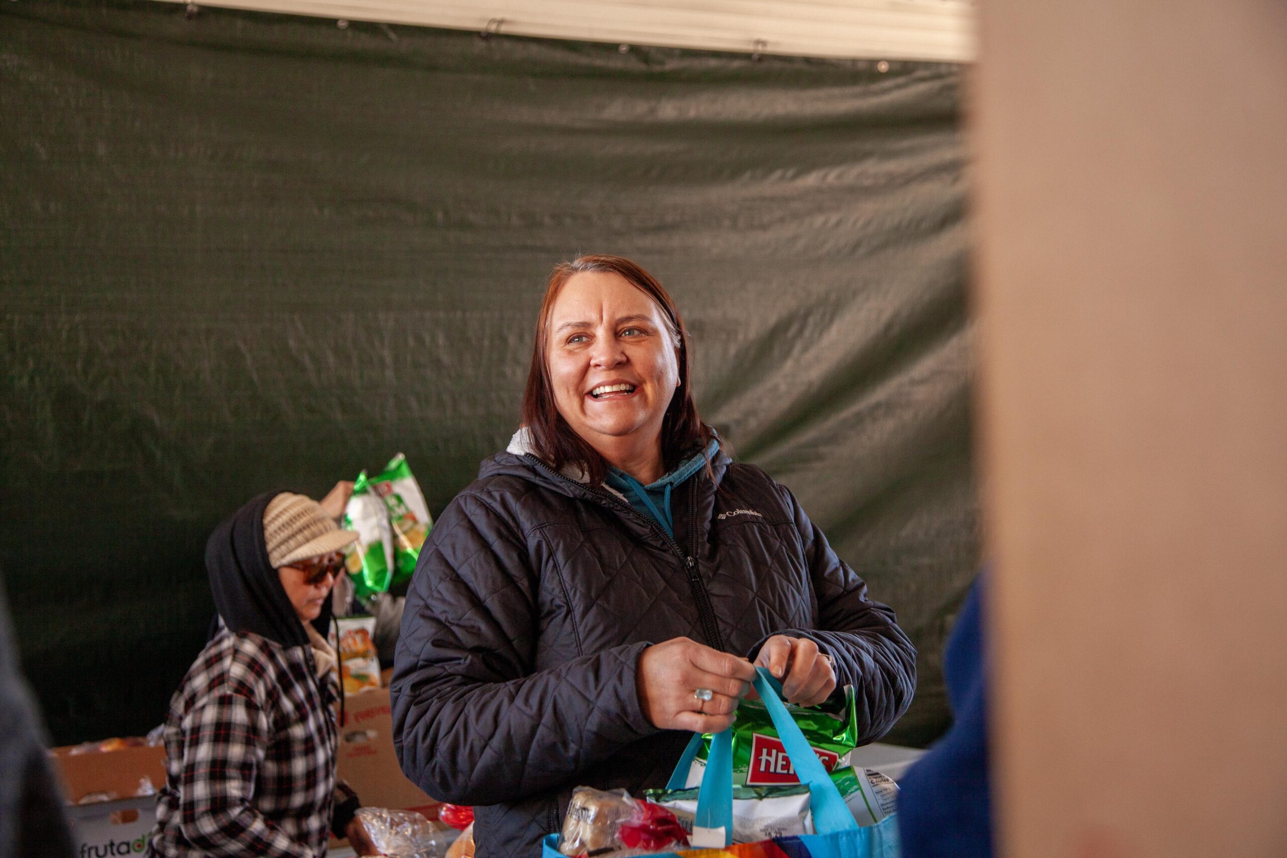 Smiling woman holding a reusable shopping bag filled with groceries at a food distribution event, with another participant in the background, set against a green tent backdrop.