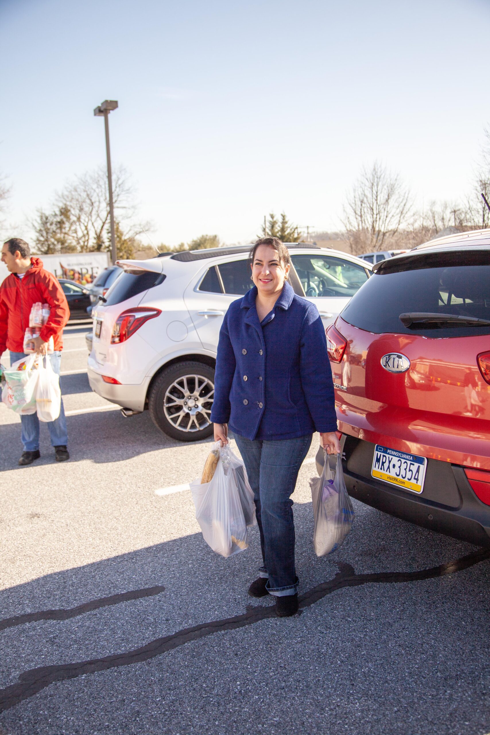 Woman smiling while carrying grocery bags in a parking lot, with cars in the background and a clear blue sky above.