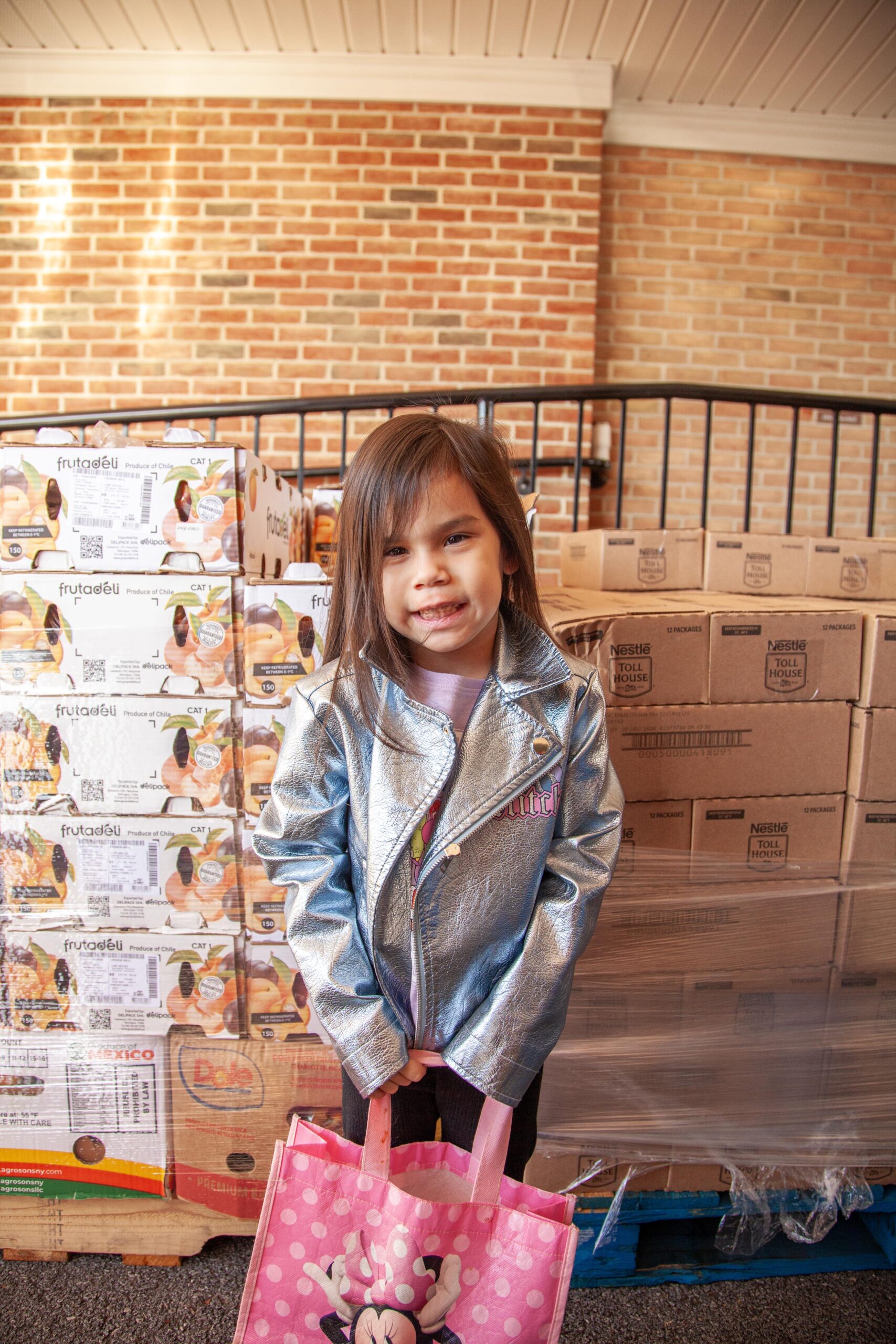 Smiling young girl wearing a shiny silver jacket and holding a pink Minnie Mouse tote bag stands in front of stacked boxes of food products in a community distribution center.