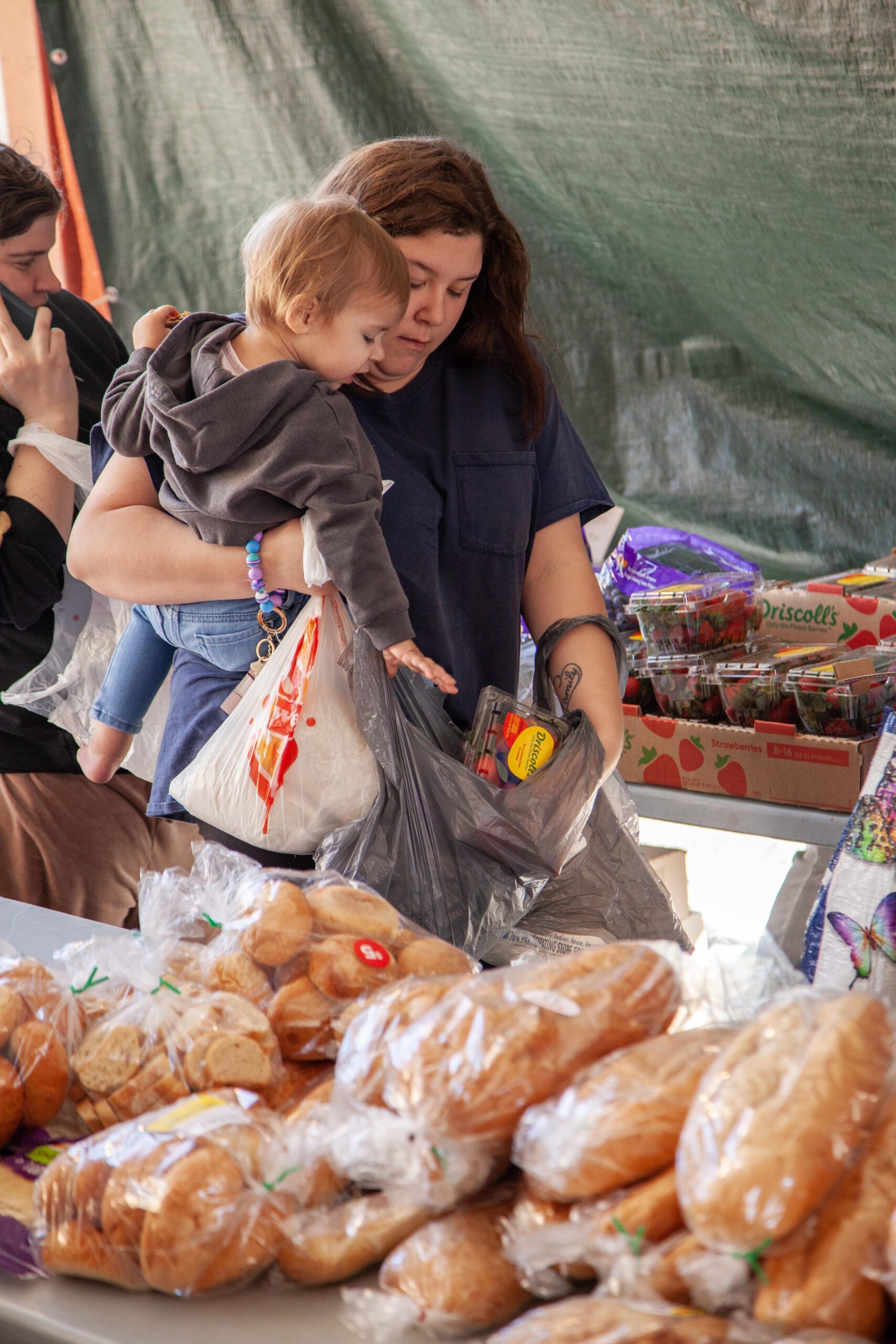 Mother shopping at a market with a toddler in her arms, surrounded by various types of fresh bread and produce, showcasing a vibrant community atmosphere.
