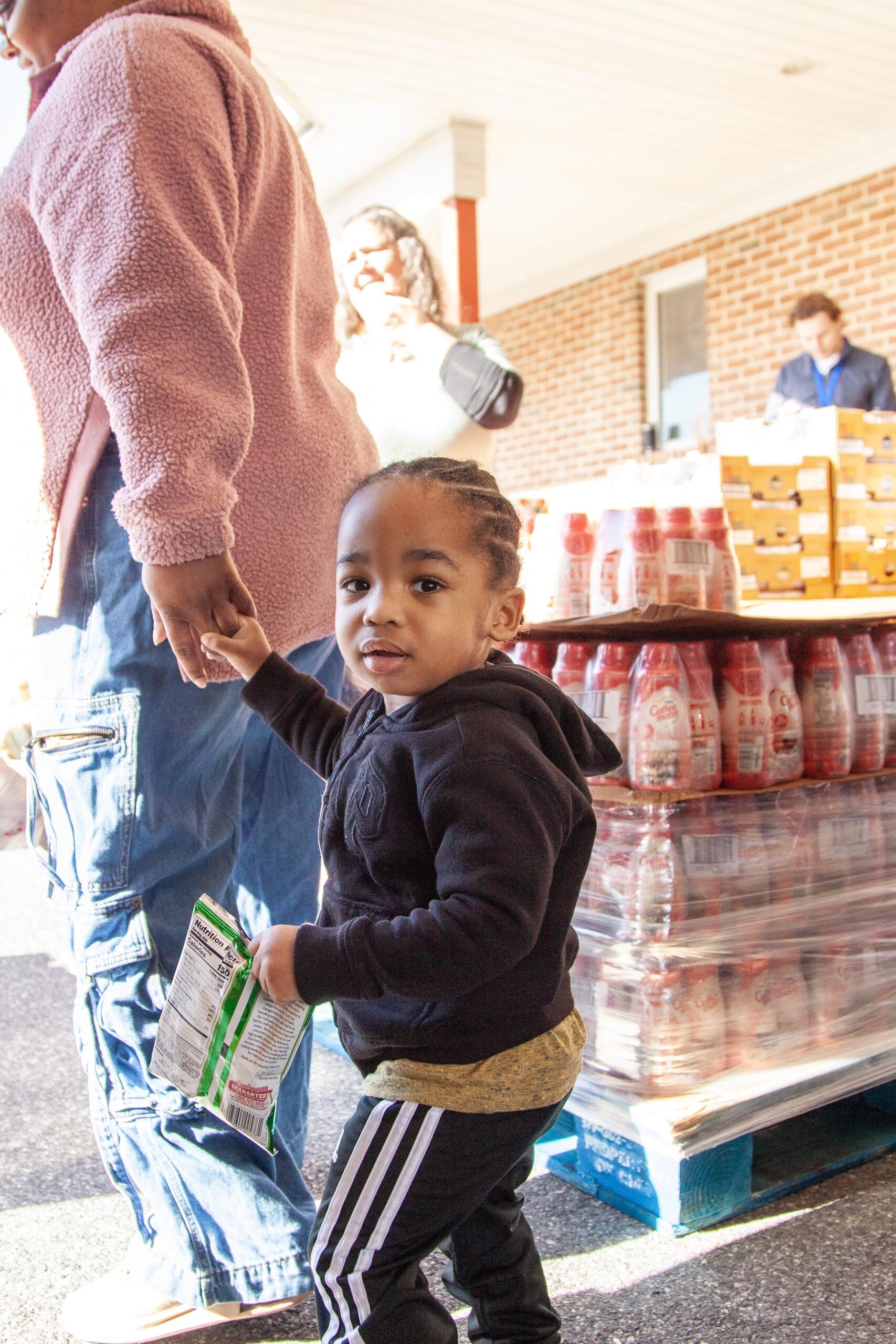 Child holding a snack while holding hands with an adult, standing in front of a food distribution area with stacked goods in the background.