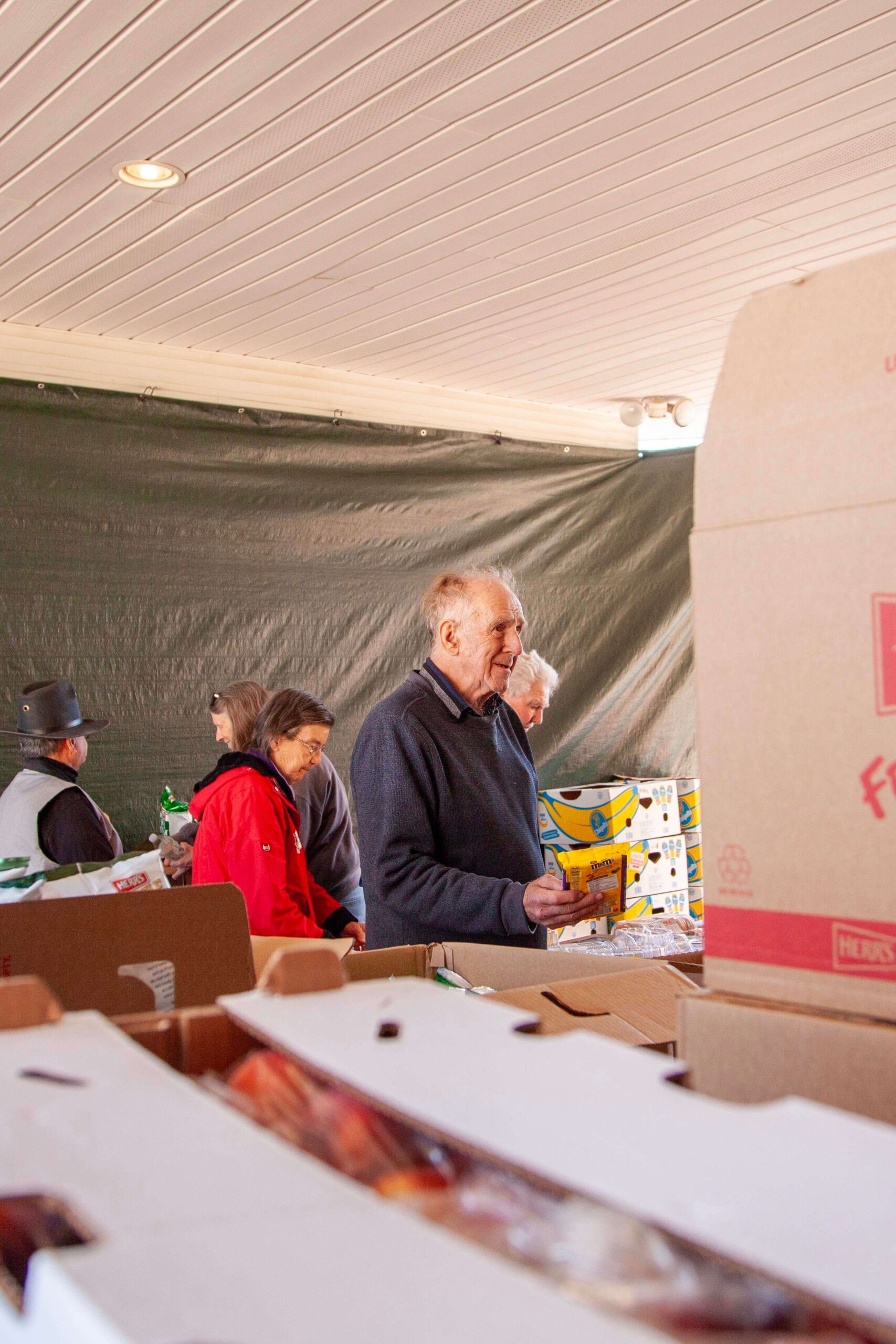 Elderly man holding a package while shopping at a community market, surrounded by boxes of food and other shoppers in a bustling indoor setting.