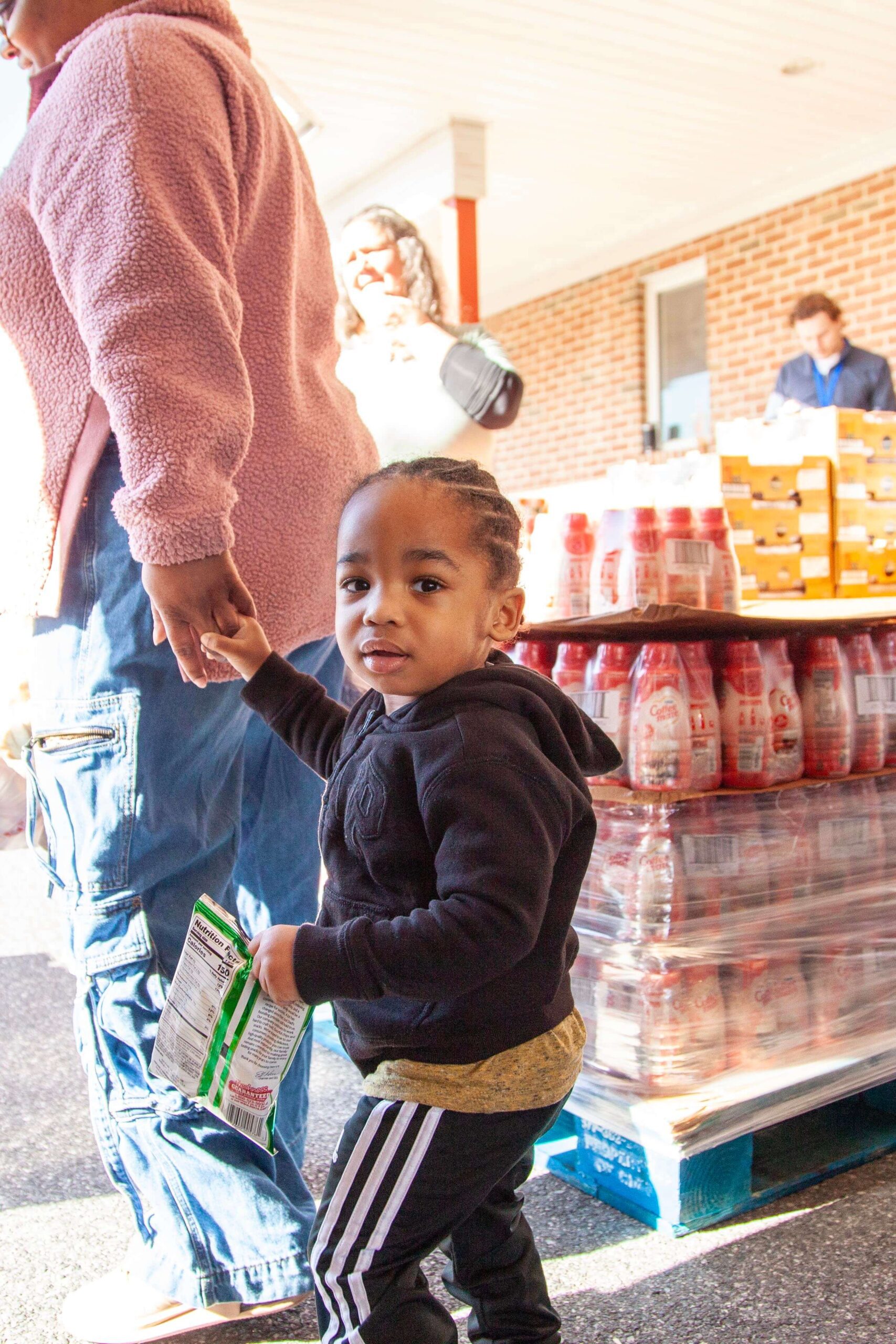 A young child in a black hoodie holds a snack bag while walking hand-in-hand with an adult wearing a pink fleece jacket. In the background, shelves stacked with food items are visible, indicating a community event or food distribution.