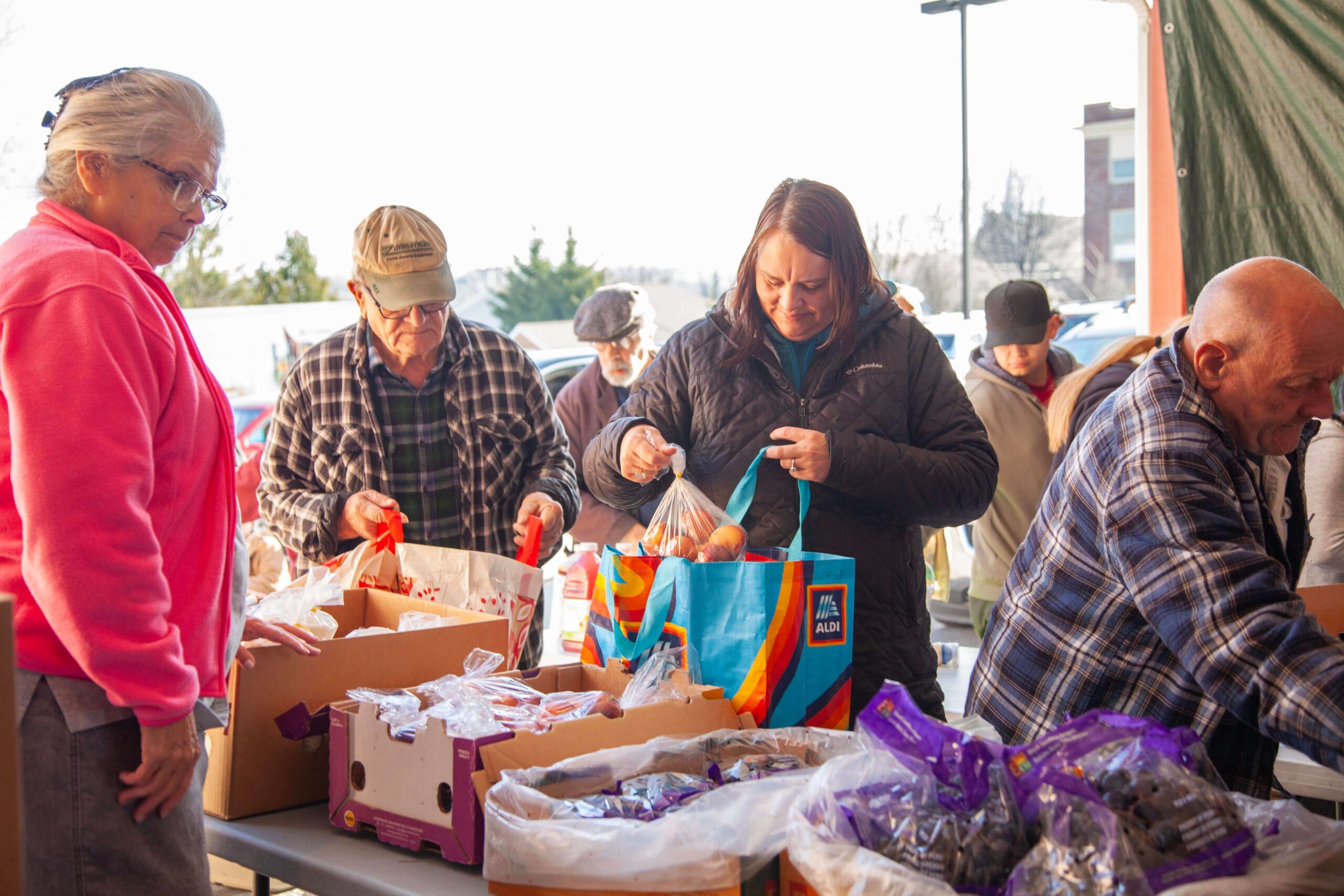 Community members gather at a food distribution event, selecting fresh produce and packaged goods from tables filled with boxes. The scene captures a diverse group of individuals, including seniors and families, engaging in the process of obtaining food resources in a supportive environment.