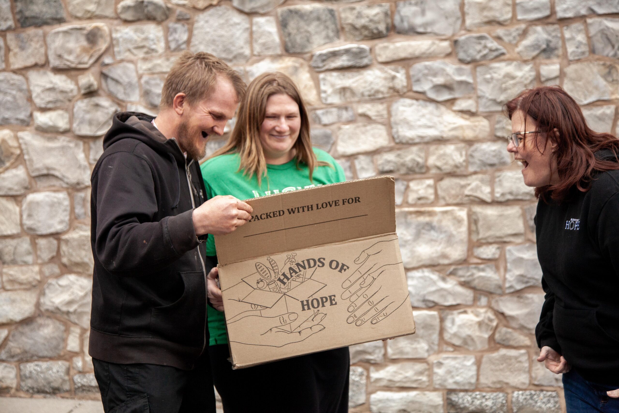 Three volunteers joyfully unbox a donation labeled "Hands of Hope," showcasing community support and engagement, against a stone wall backdrop.