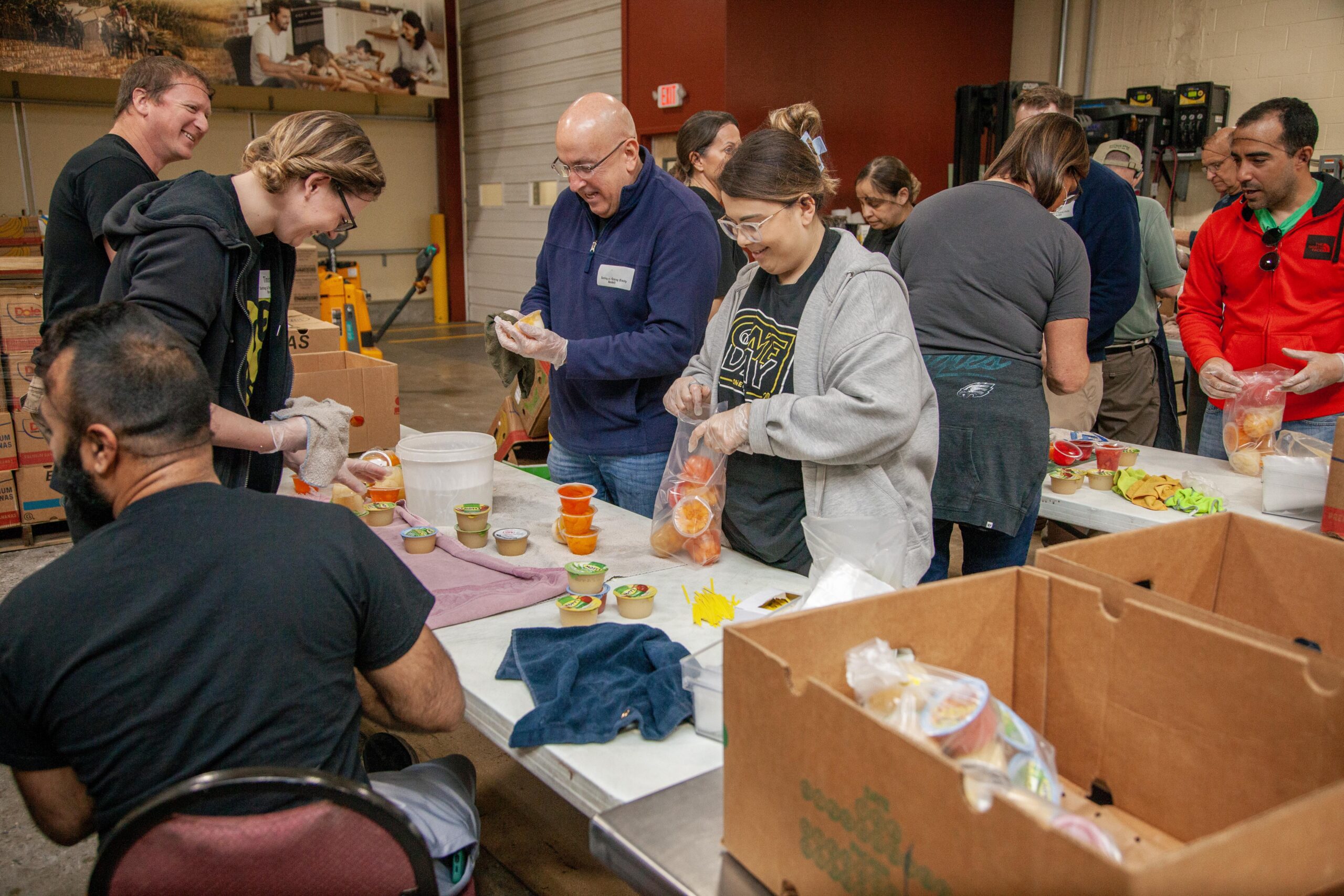Volunteers packing food items in a community kitchen, engaging in teamwork to prepare meals for those in need. The scene shows diverse individuals collaborating at tables filled with food containers and supplies, highlighting community service and support.