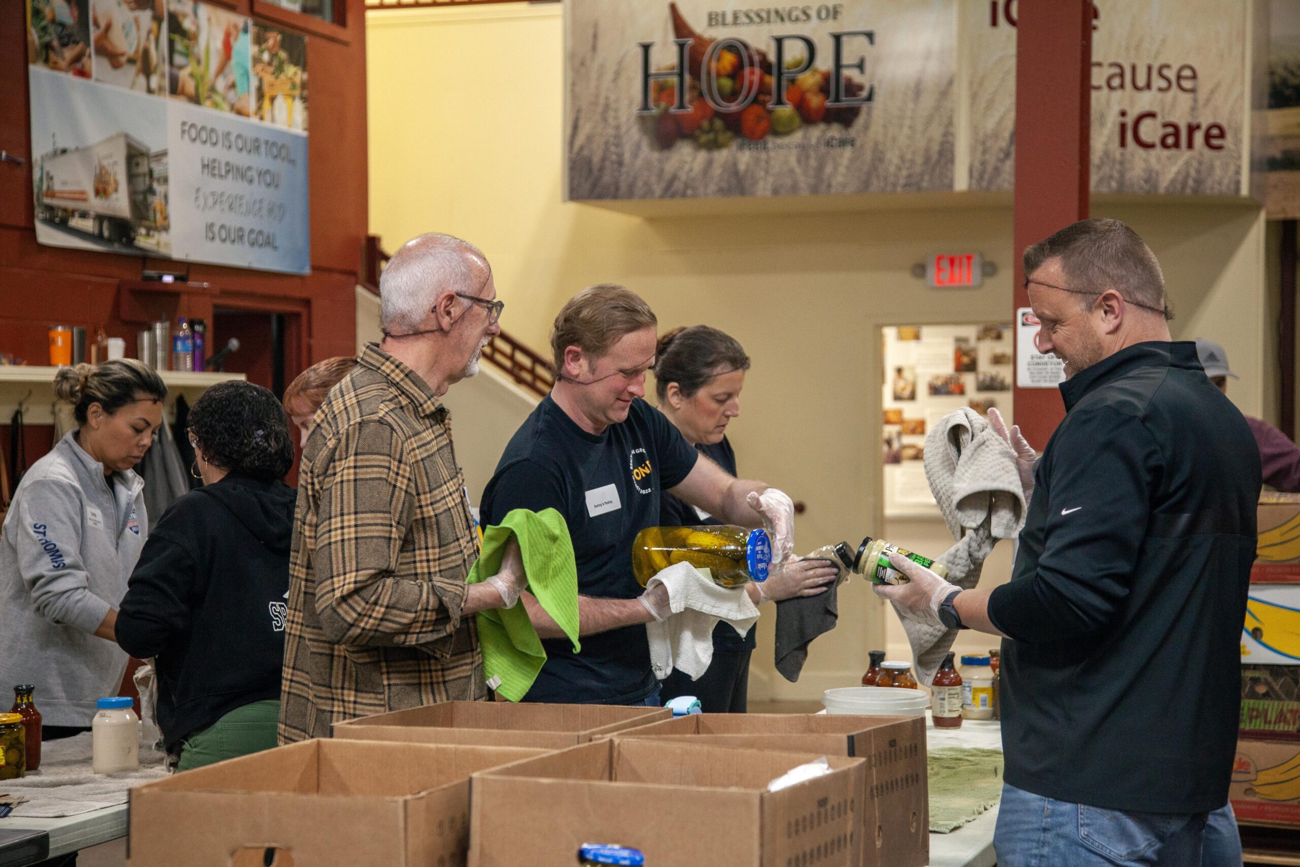 Volunteers working together at a food distribution center, cleaning and preparing items for packing into boxes. The backdrop features a sign that reads "Blessings of Hope," emphasizing the mission to help those in need.