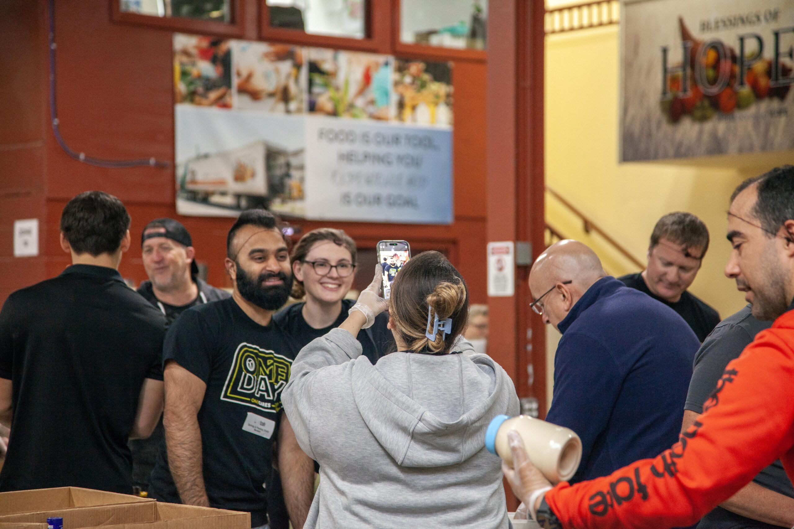 Volunteers at a community food distribution event, smiling and engaging with each other while one person takes a selfie. The background features banners promoting food assistance and community support initiatives.