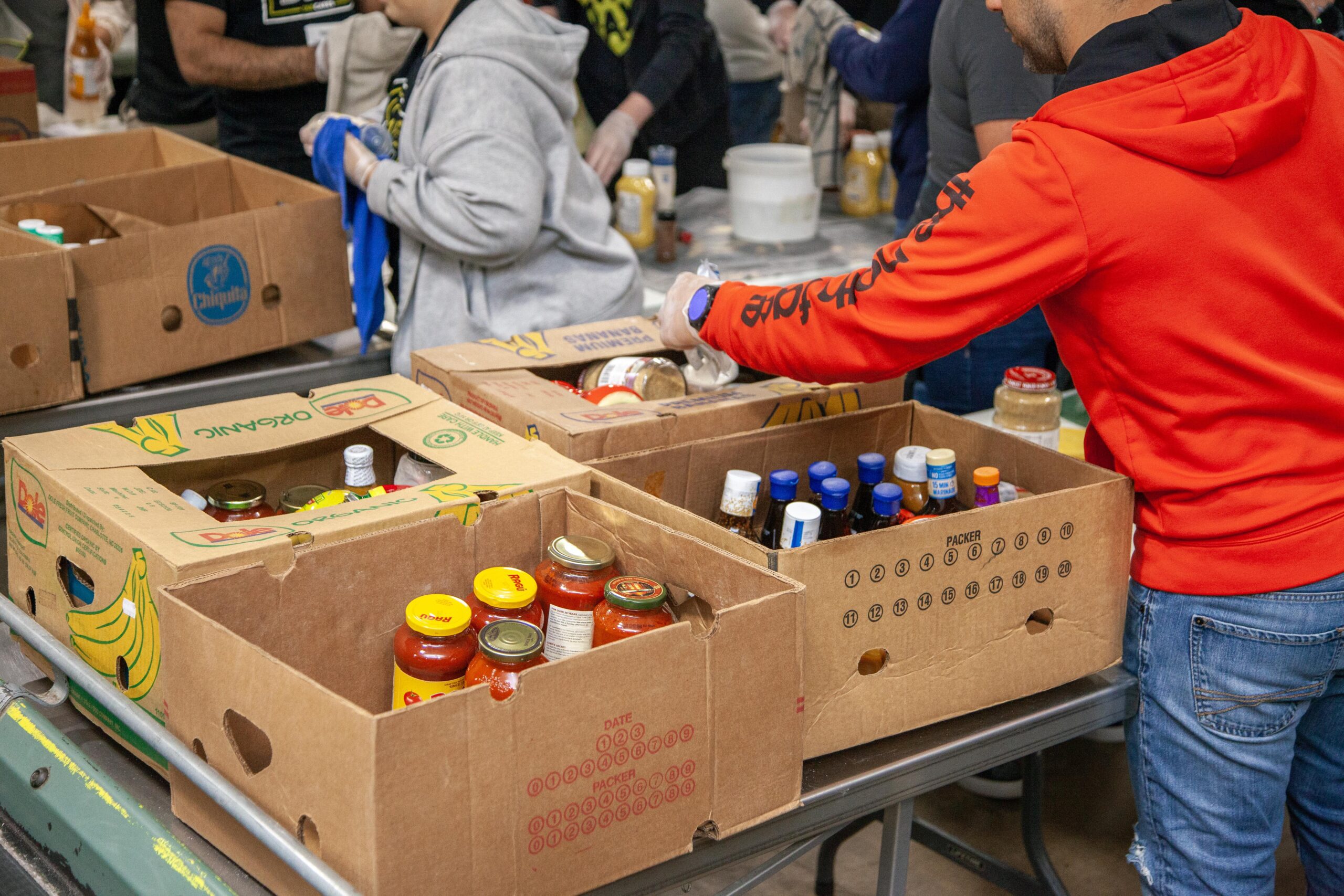 Volunteers organizing food items in cardboard boxes at a community food distribution event, with various sauces and condiments visible.