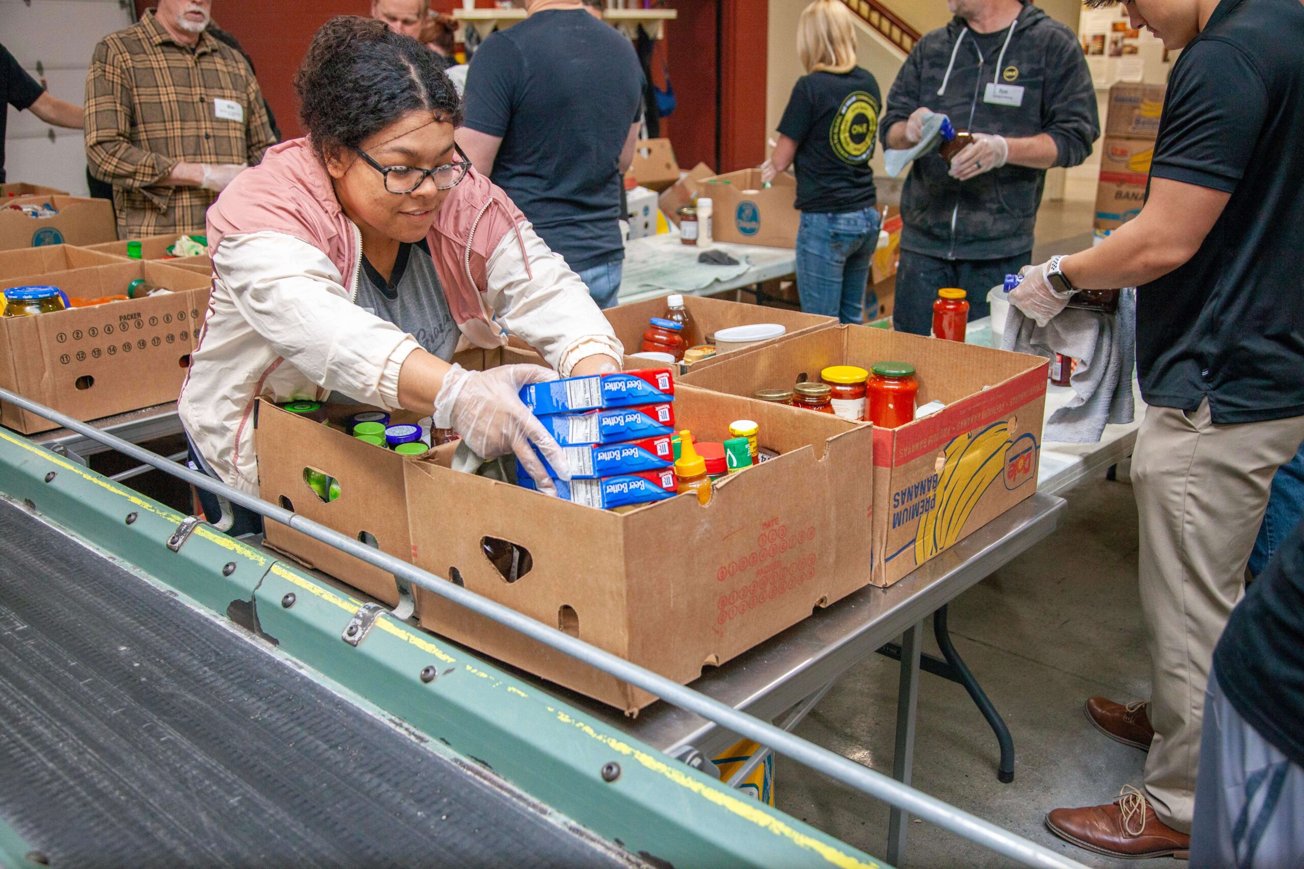 Volunteer sorting food items in cardboard boxes at a community food bank, with various jars and containers visible on a table.