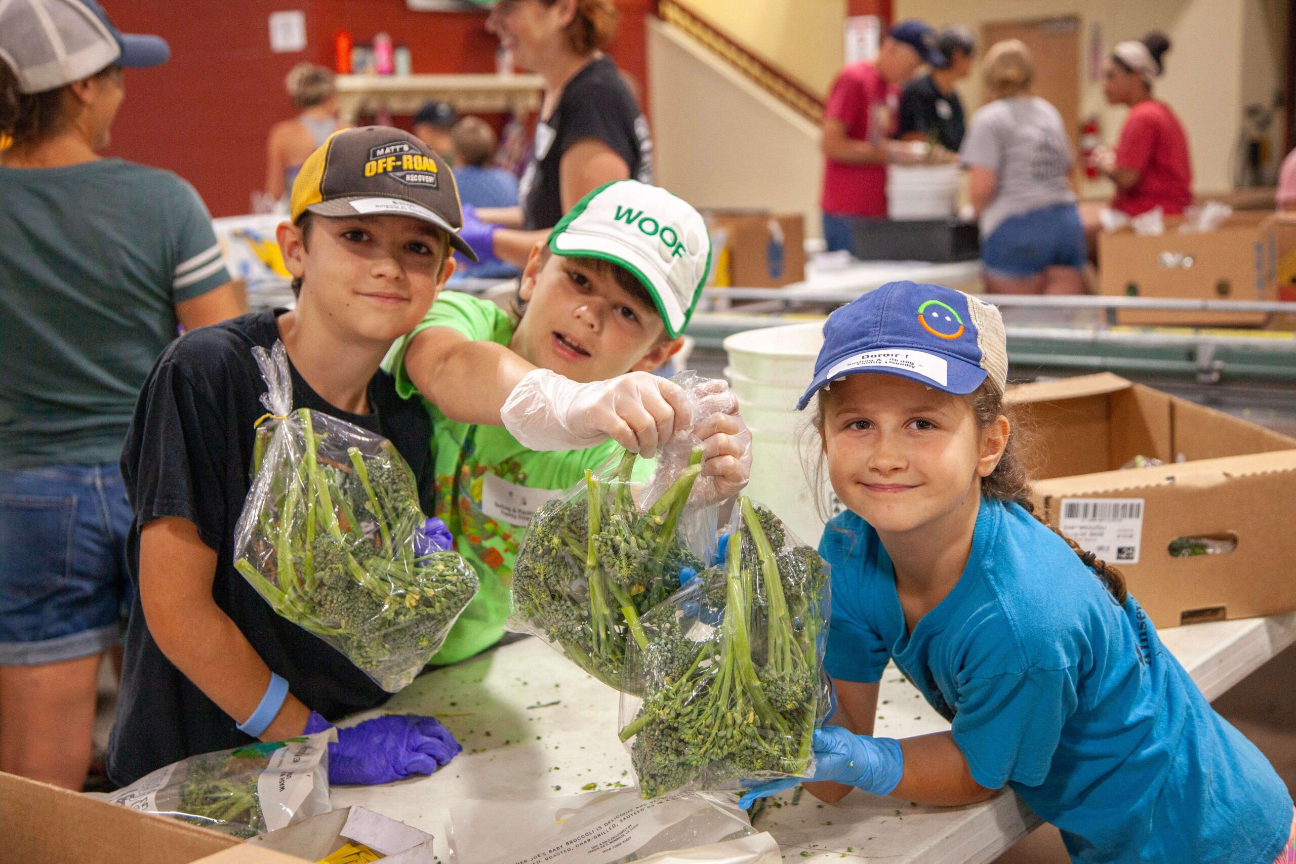 Children volunteering in a food packing event, smiling and holding bags of fresh broccoli, showcasing community service and teamwork in action.