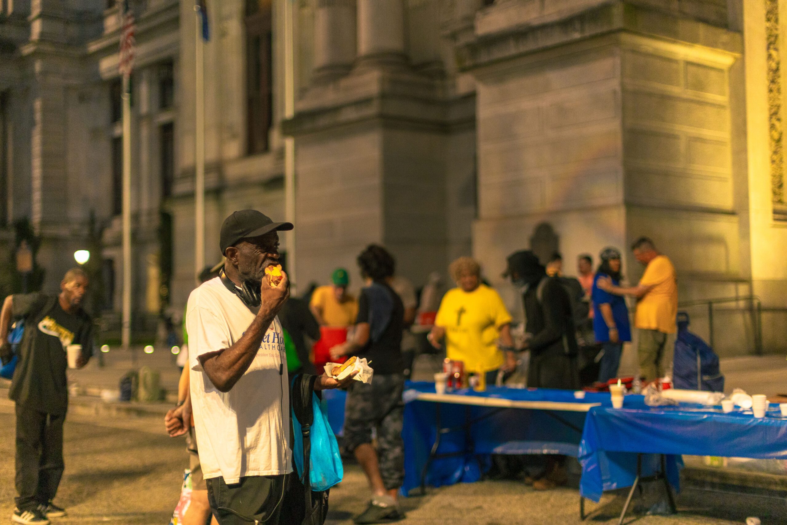 A man enjoying food stands in front of a gathering of people in a nighttime urban setting, with tables set up for a community event. Various individuals, some wearing colorful shirts, are seen engaging in conversation and serving food. The scene highlights community support and outreach efforts.