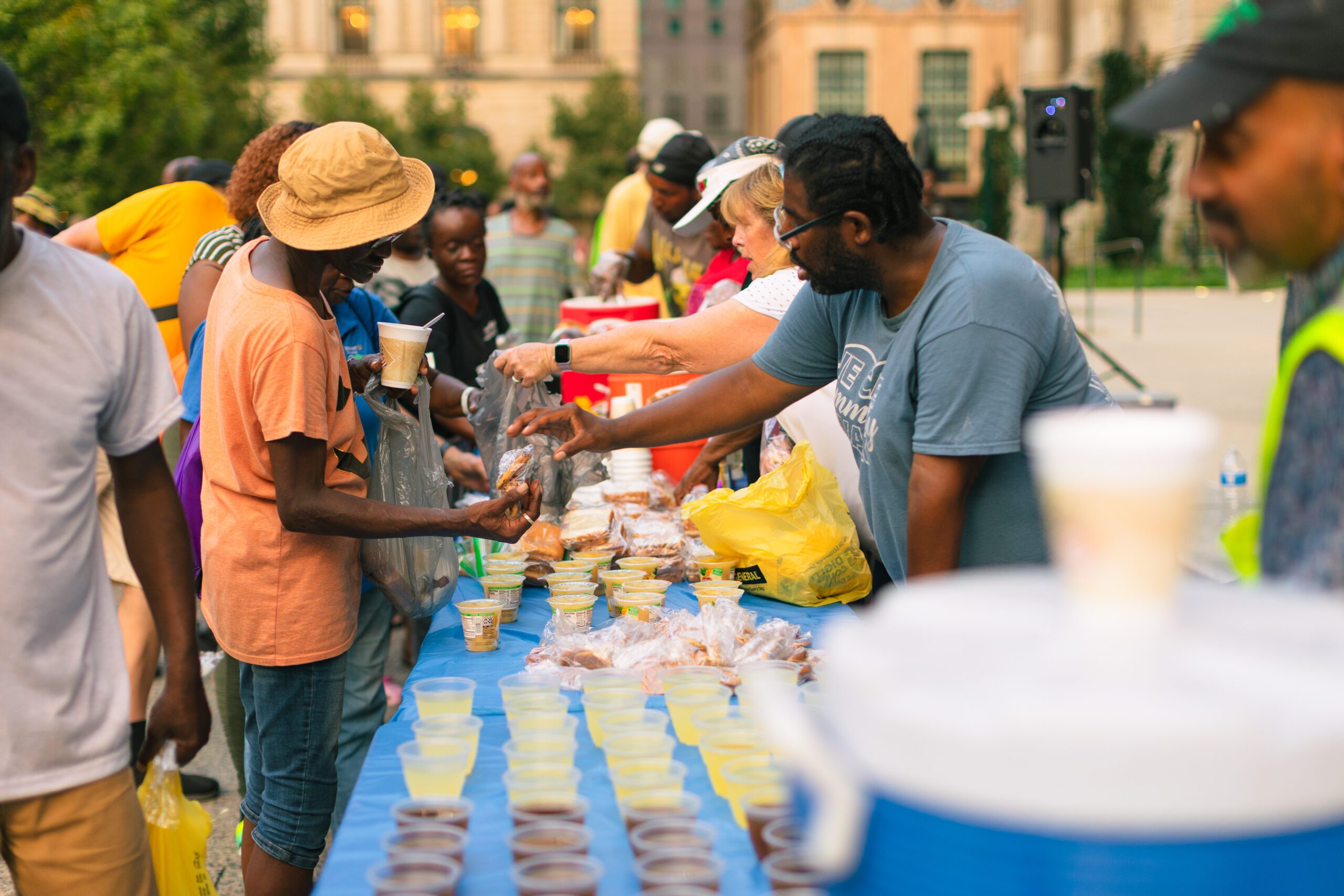 Community members gather at a food distribution event, interacting with volunteers as they receive meals and beverages from a long table filled with food items in a vibrant outdoor setting.