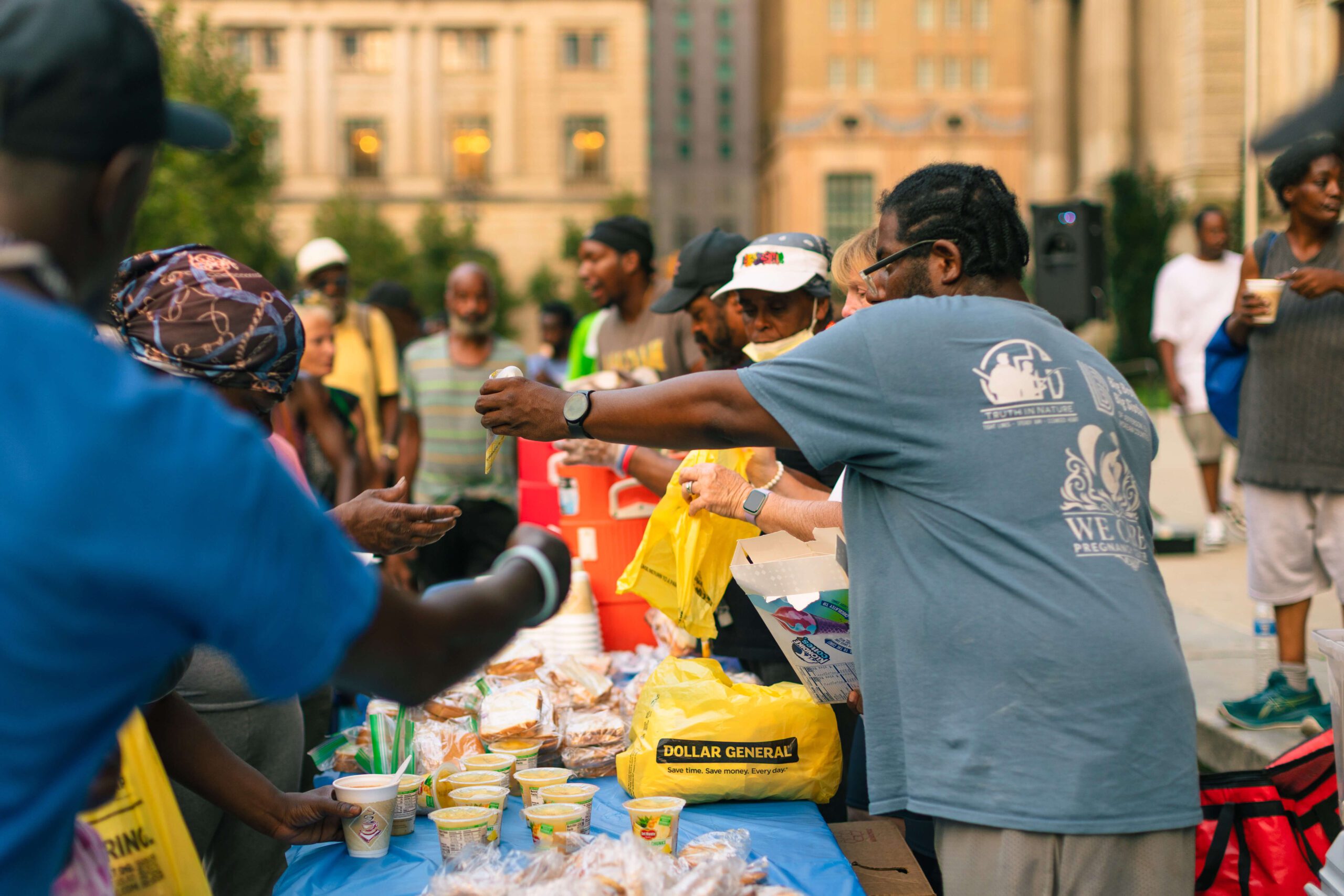 Community members gather around a table filled with food and drinks during a local outreach event, exchanging items and enjoying a sense of togetherness.
