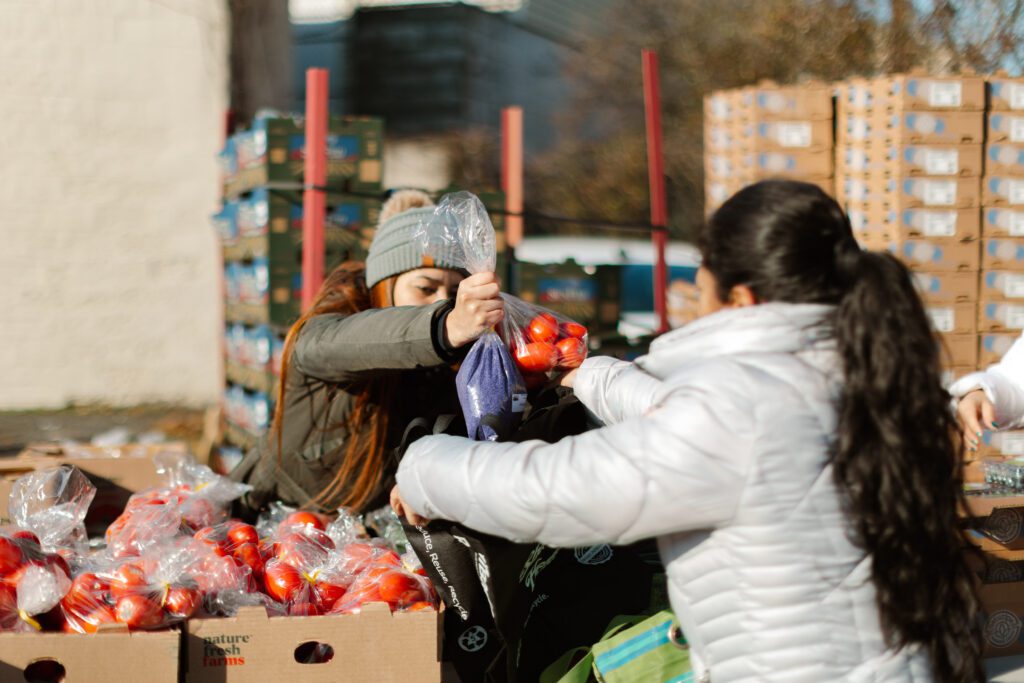 Two women participate in a community food distribution event, exchanging bags of apples at an outdoor market. Boxes of fresh produce are stacked in the background, highlighting the initiative to provide healthy food options.