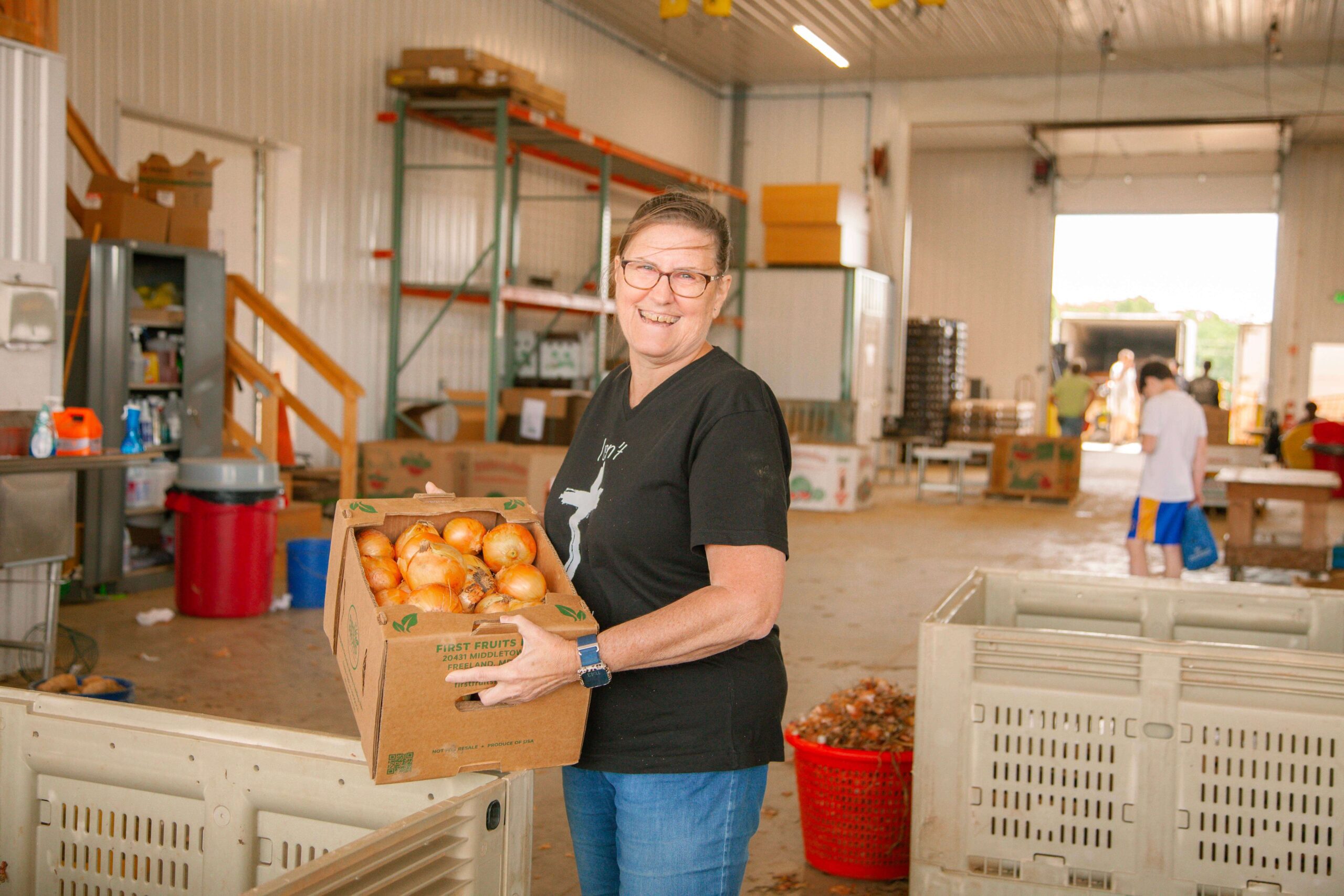 Smiling woman holding a box of fresh onions in a produce warehouse, surrounded by crates and storage areas, showcasing community farming efforts and local food distribution.