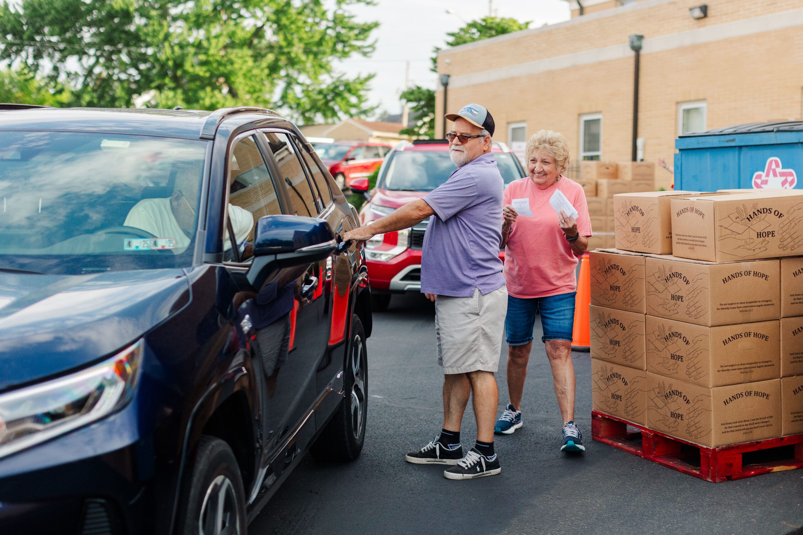 Volunteers assisting at a food distribution event, with a man opening a car door and a woman holding tickets in front of stacked boxes labeled "Hands of Hope." The scene captures community support and charitable efforts.