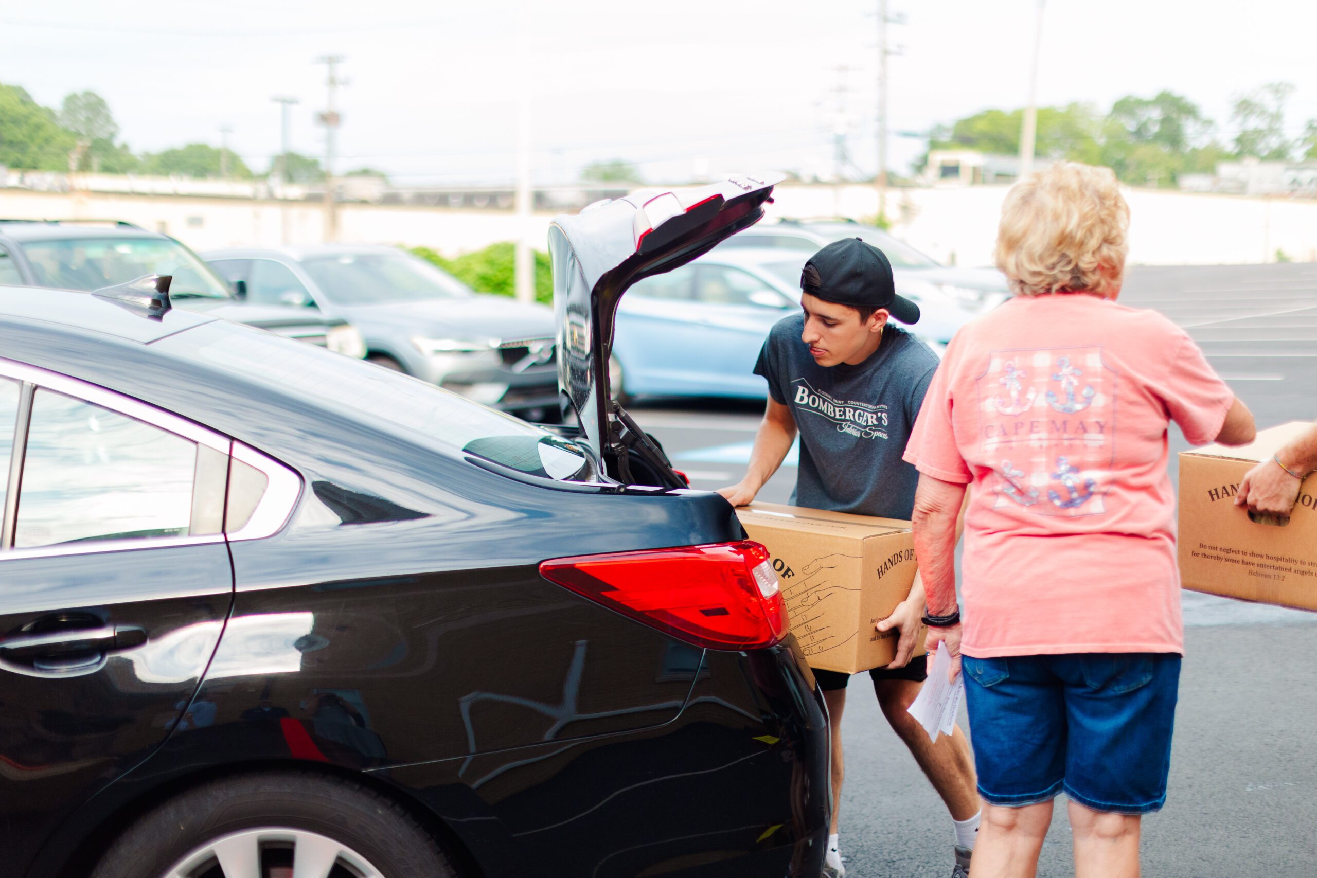 A young man loading boxes into the trunk of a black car while an older woman holds another box, in a parking lot setting.