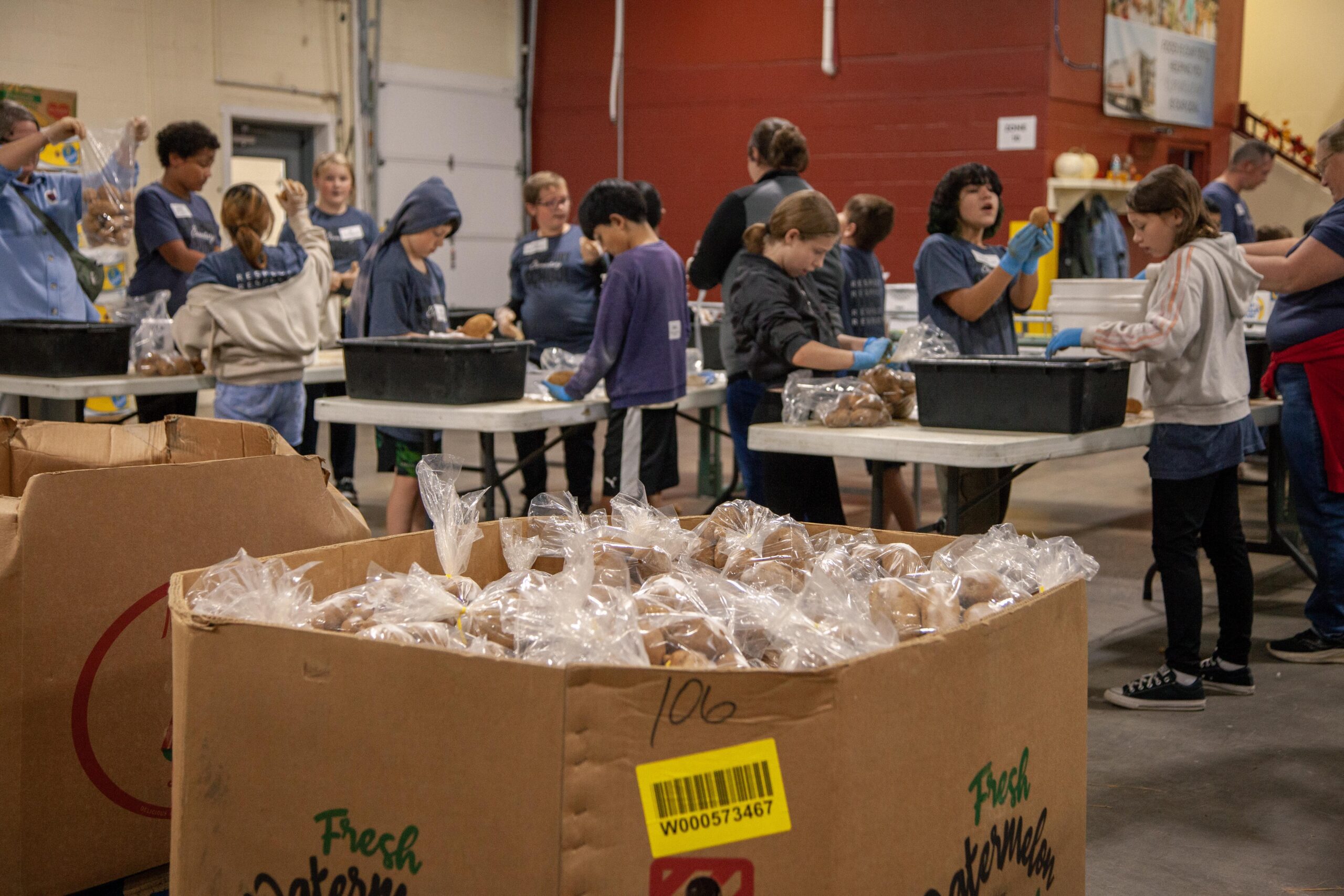 Volunteers packing fresh baked goods in a food distribution center, with children and adults working together at tables, surrounded by boxes filled with packaged items.