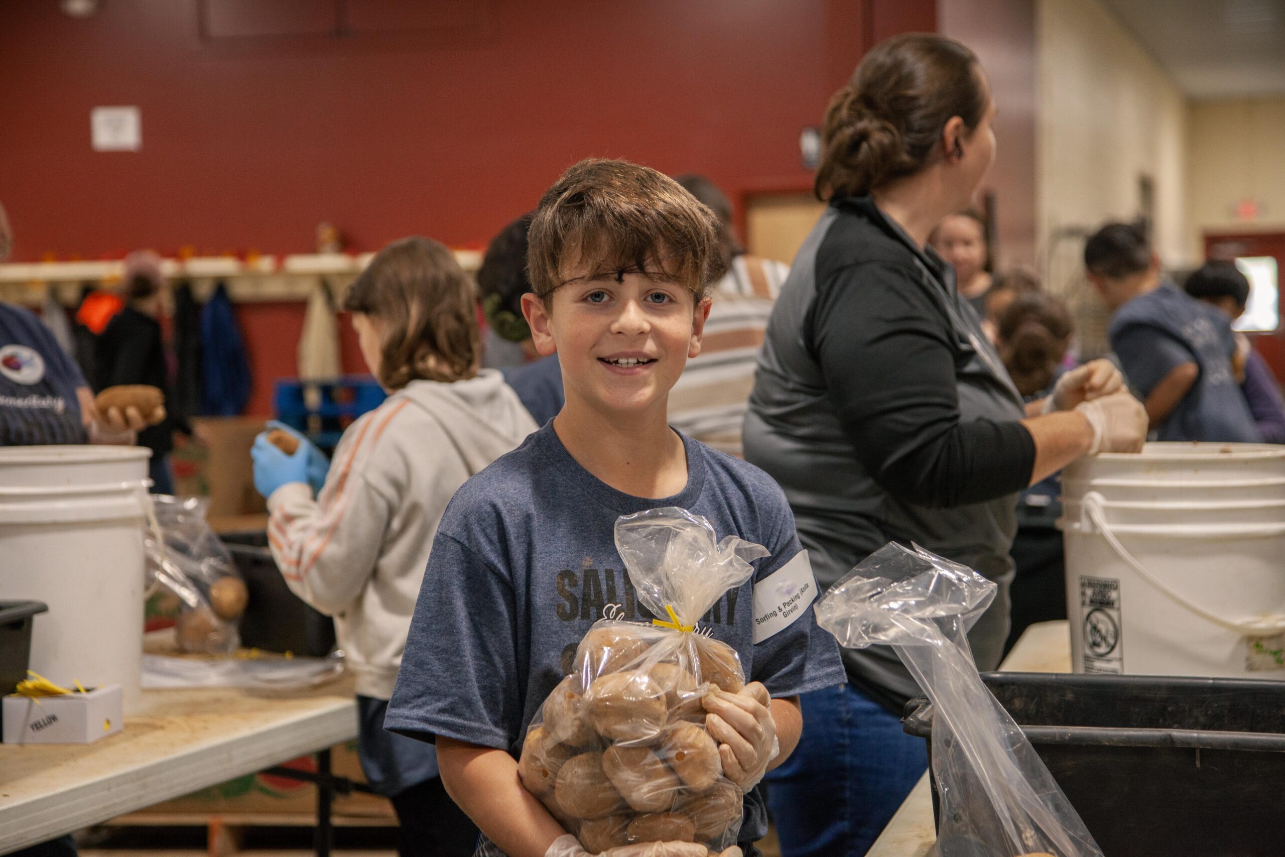 Young boy smiling while holding a bag of potatoes at a food distribution event, surrounded by volunteers packing food in a community center.