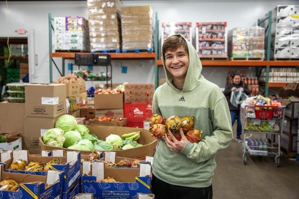 Teenager in a green hoodie smiles while holding fresh dragon fruit in a grocery store filled with various fruits and vegetables. Boxes of produce are stacked around him, showcasing a vibrant selection of healthy food options.